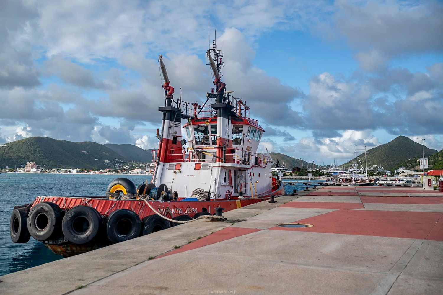 A red and white tugboat docked at a harbor pier with tire bumpers, mountains and cloudy sky in the background.