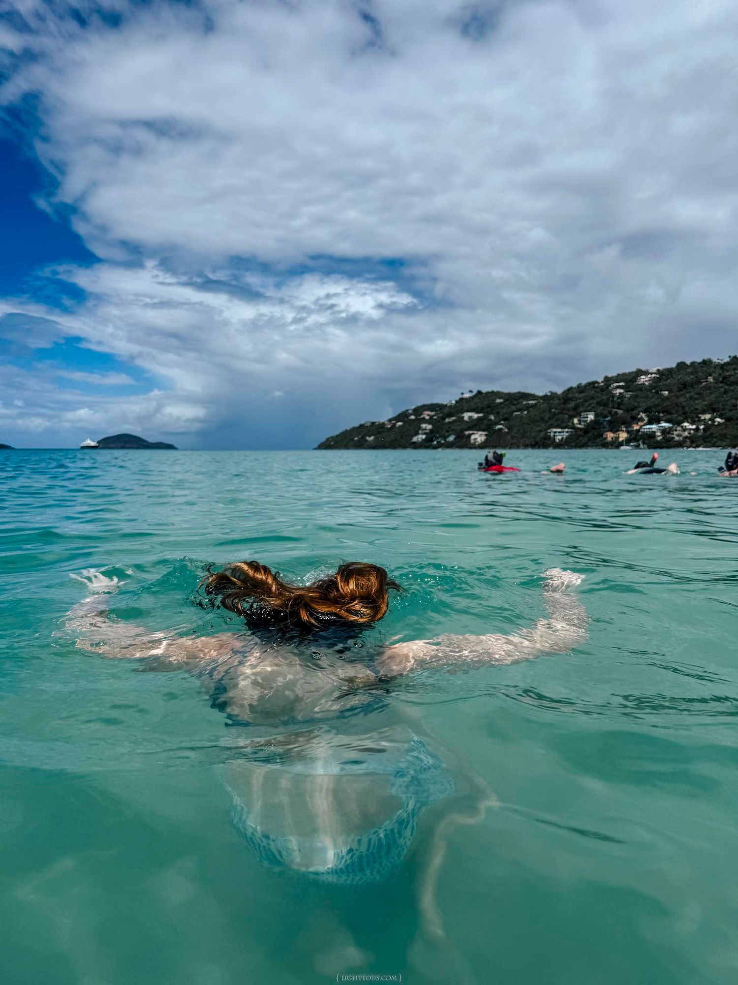 Woman swimming in clear turquoise Caribbean waters with lush green hillside and sailboats in background.