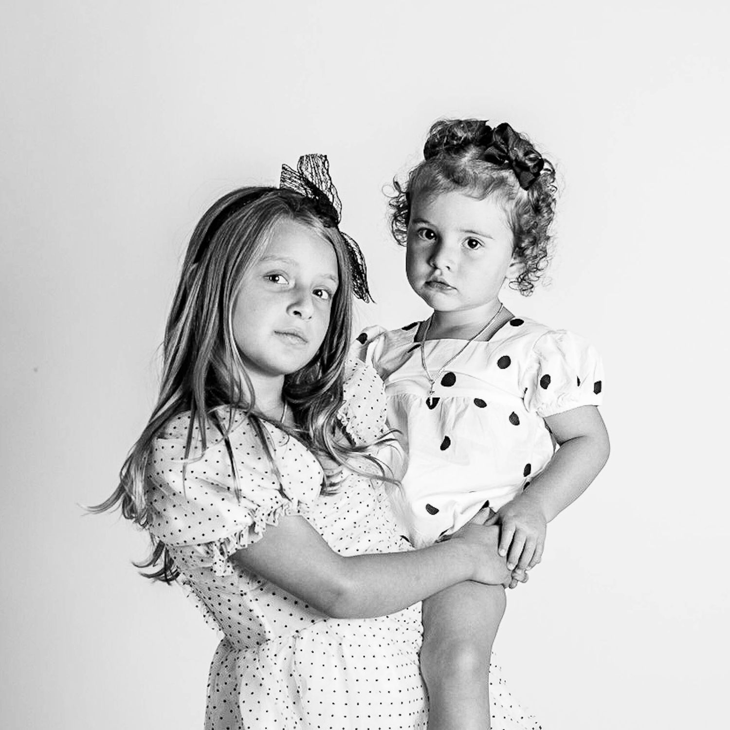 Black and white portrait of two young sisters in polka dot dresses, older girl holding younger toddler sister.