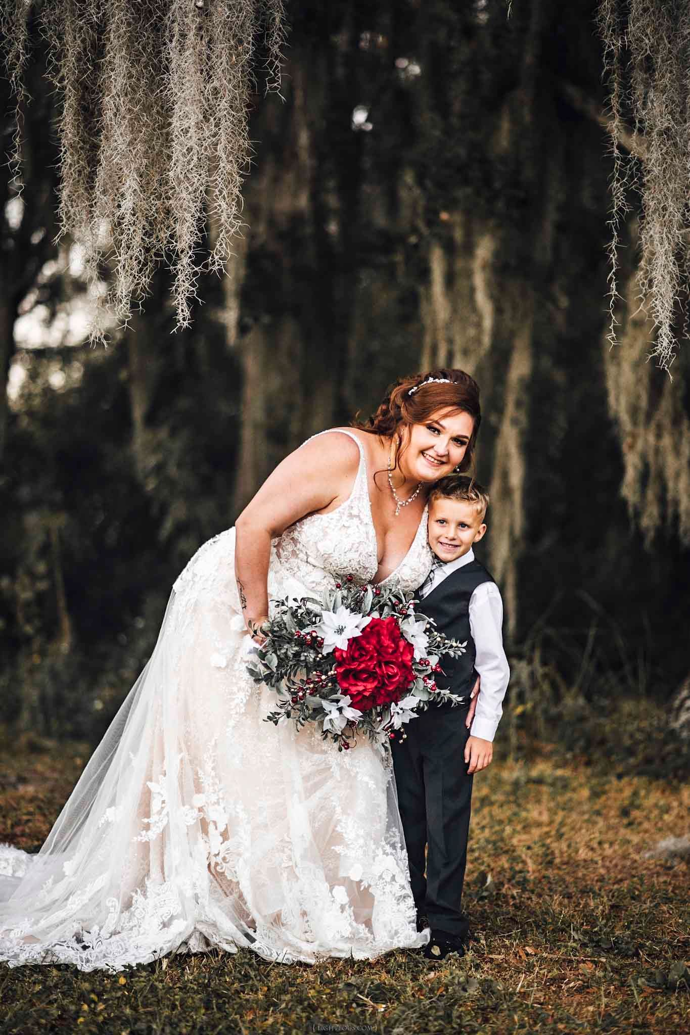 Bride in lace gown holds red bouquet beside young boy in suit under Spanish moss-draped trees.