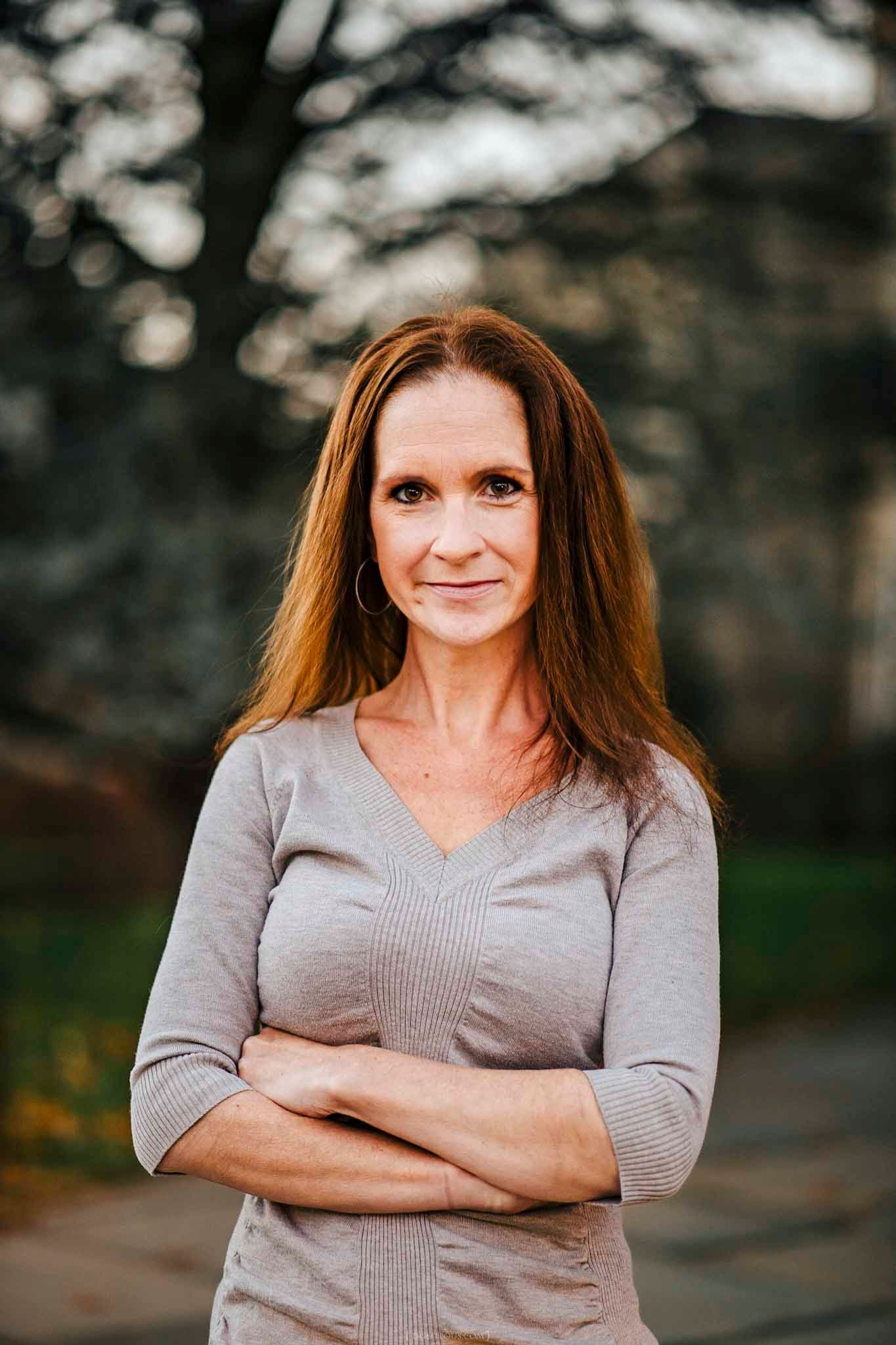 A confident woman with auburn hair stands outdoors with arms crossed, wearing a gray top, with trees in the background.