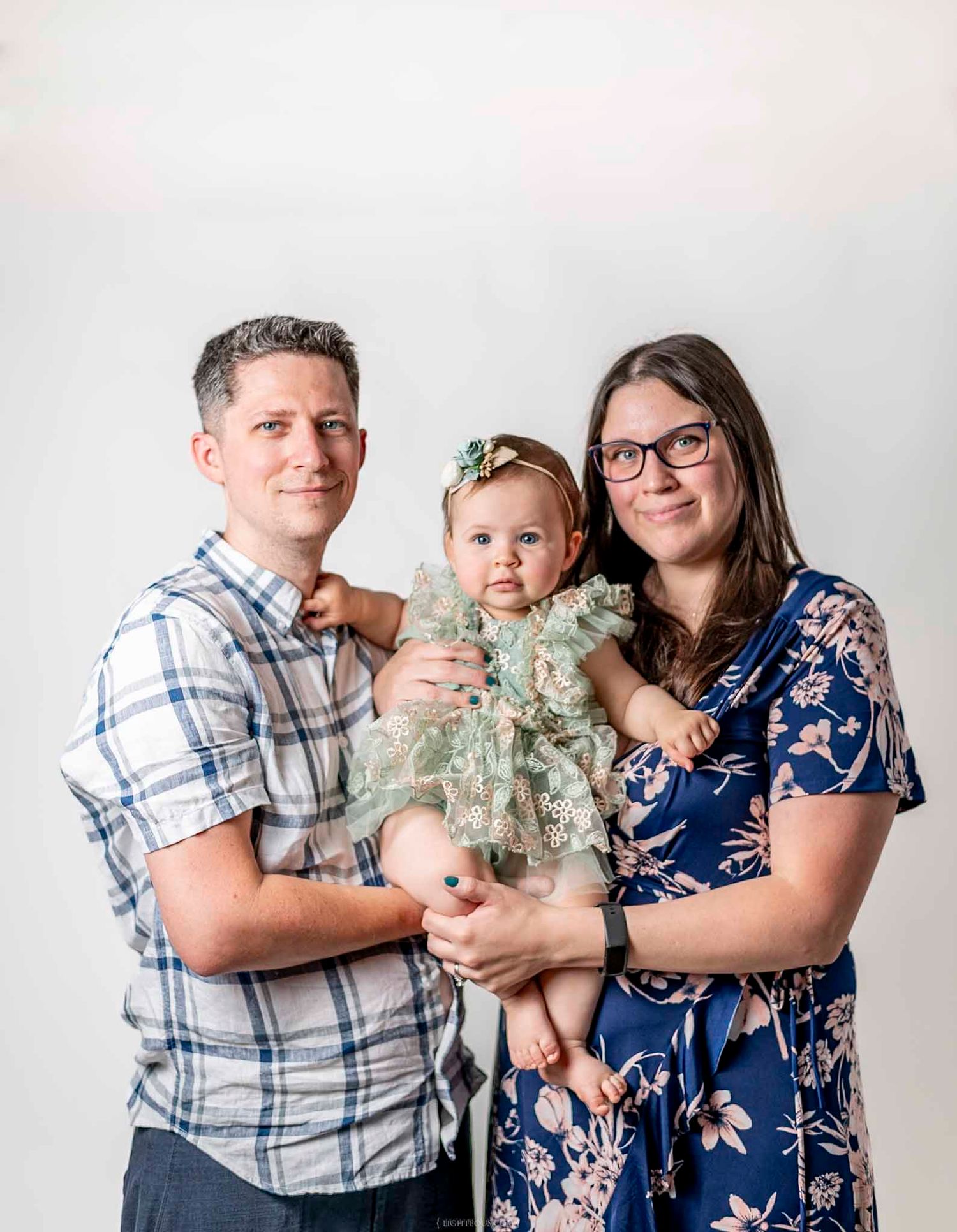 A smiling couple holds their baby girl in a floral outfit during a professional family portrait session.