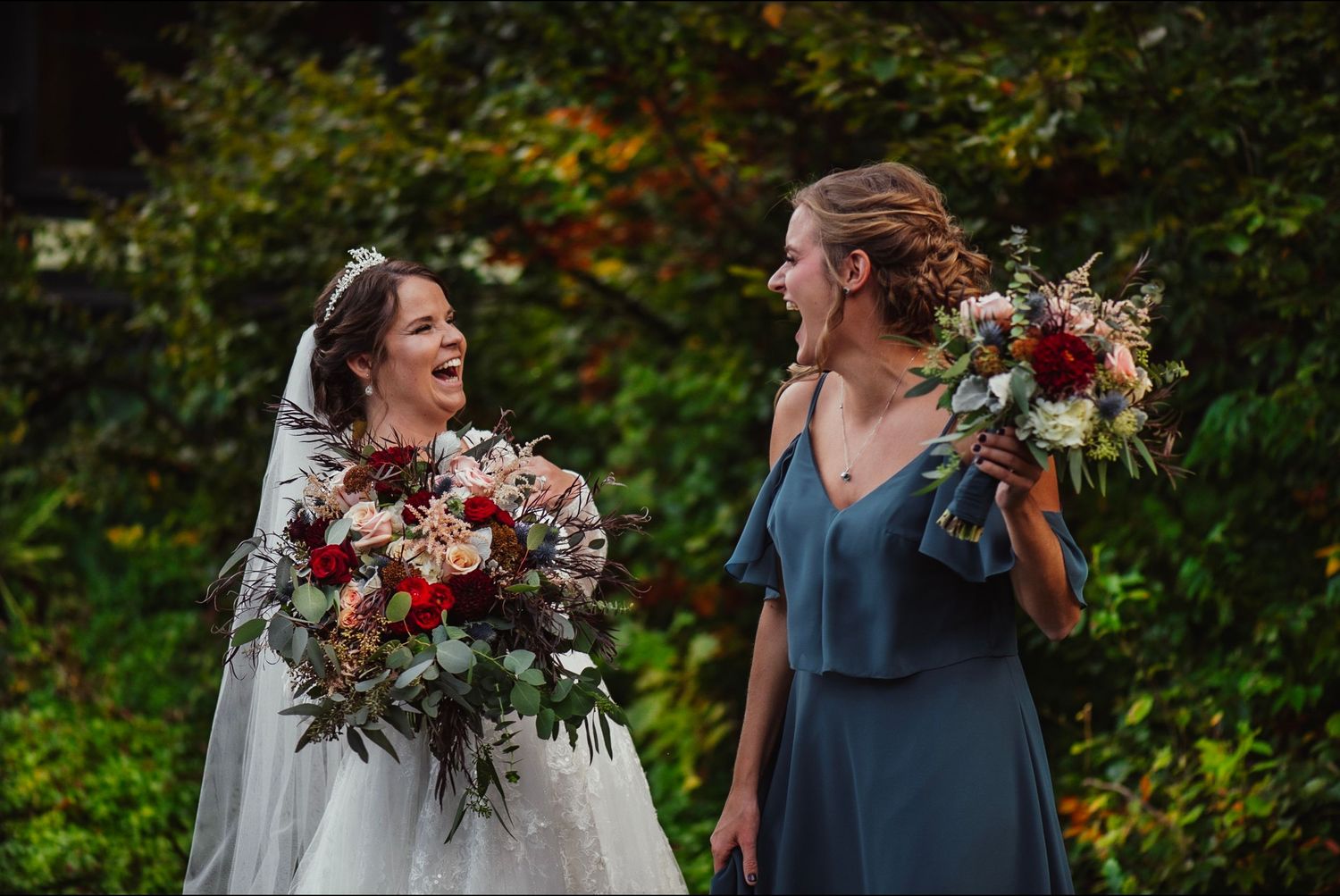 Laughing bride in white gown and bridesmaid in blue dress holding bouquets outdoors at wedding.