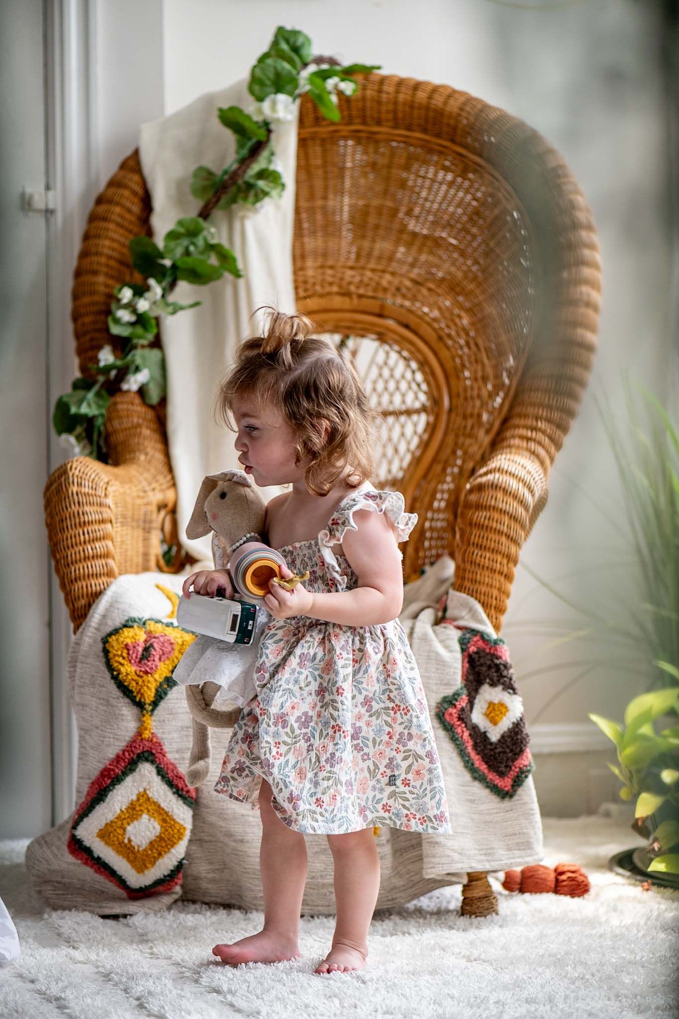 A toddler in a floral dress explores near a decorative wicker chair with colorful accents.