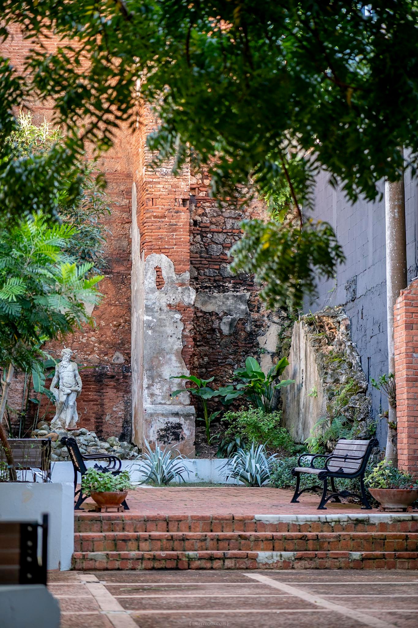 Historic courtyard with brick walls, lush greenery, classical statue, and iron benches in an old colonial garden.
