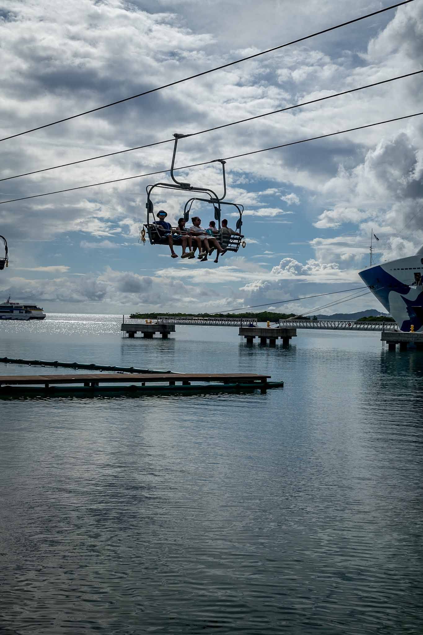 Silhouettes of chairlift riders glide over calm waters with cloudy skies and mountains in the background.