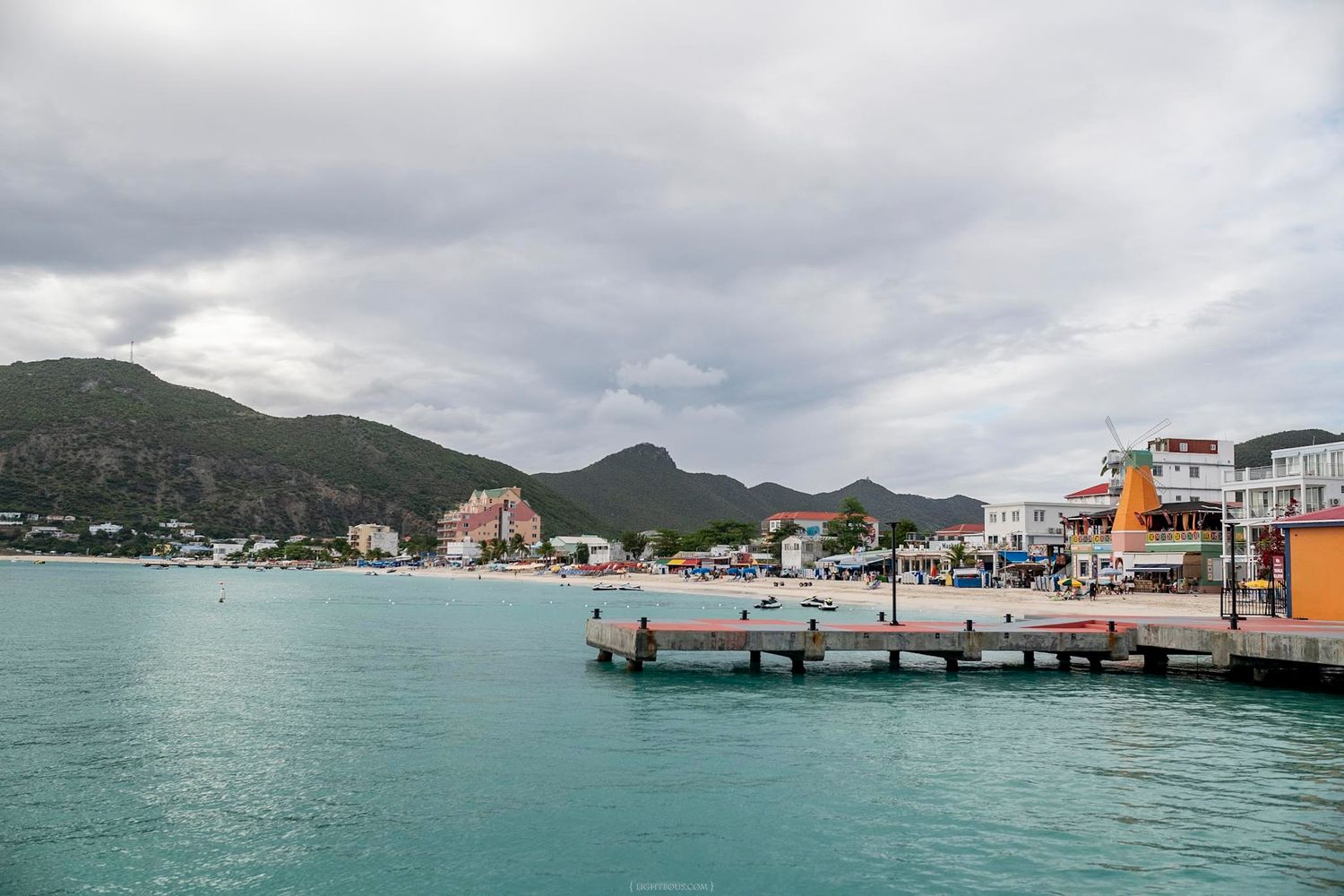 A scenic Caribbean coastal town with turquoise waters, a wooden pier, colorful buildings, and lush green mountains.