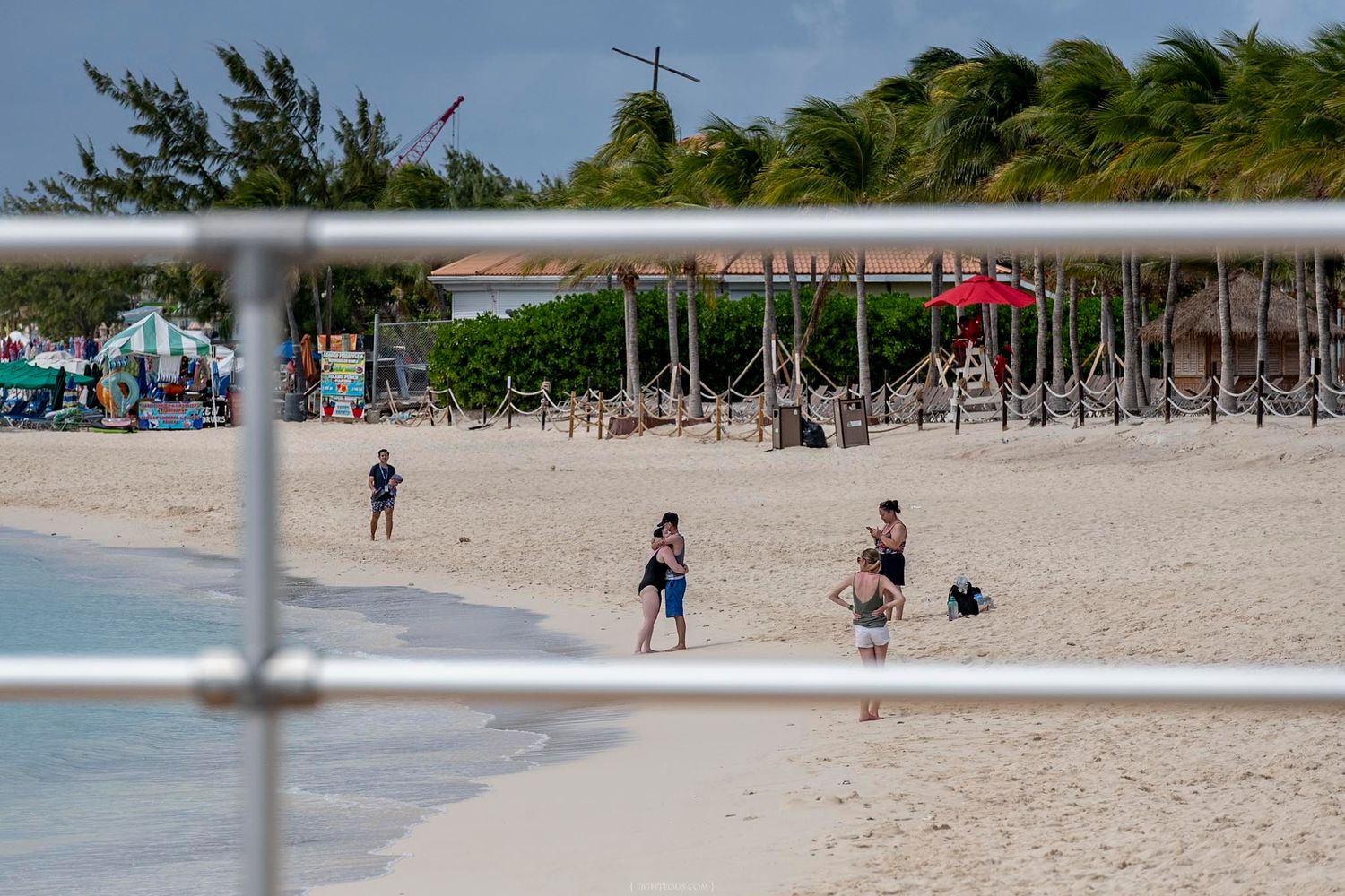 Tourists enjoying a tropical white sand beach with palm trees and colorful beach huts viewed through a metal railing.