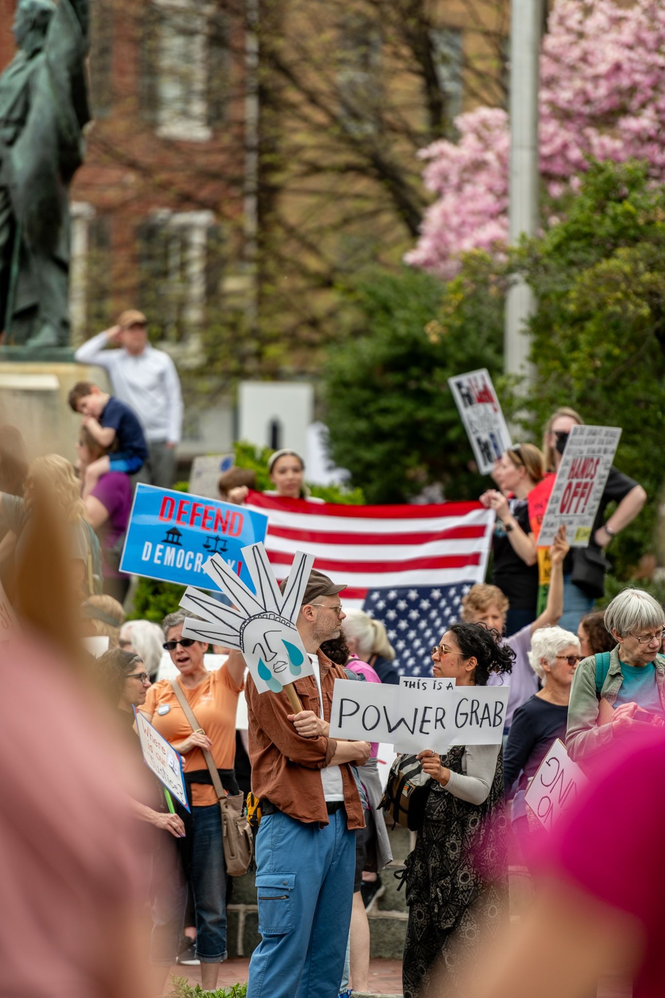 Protesters with signs and American flags at a springtime demonstration with cherry blossoms.