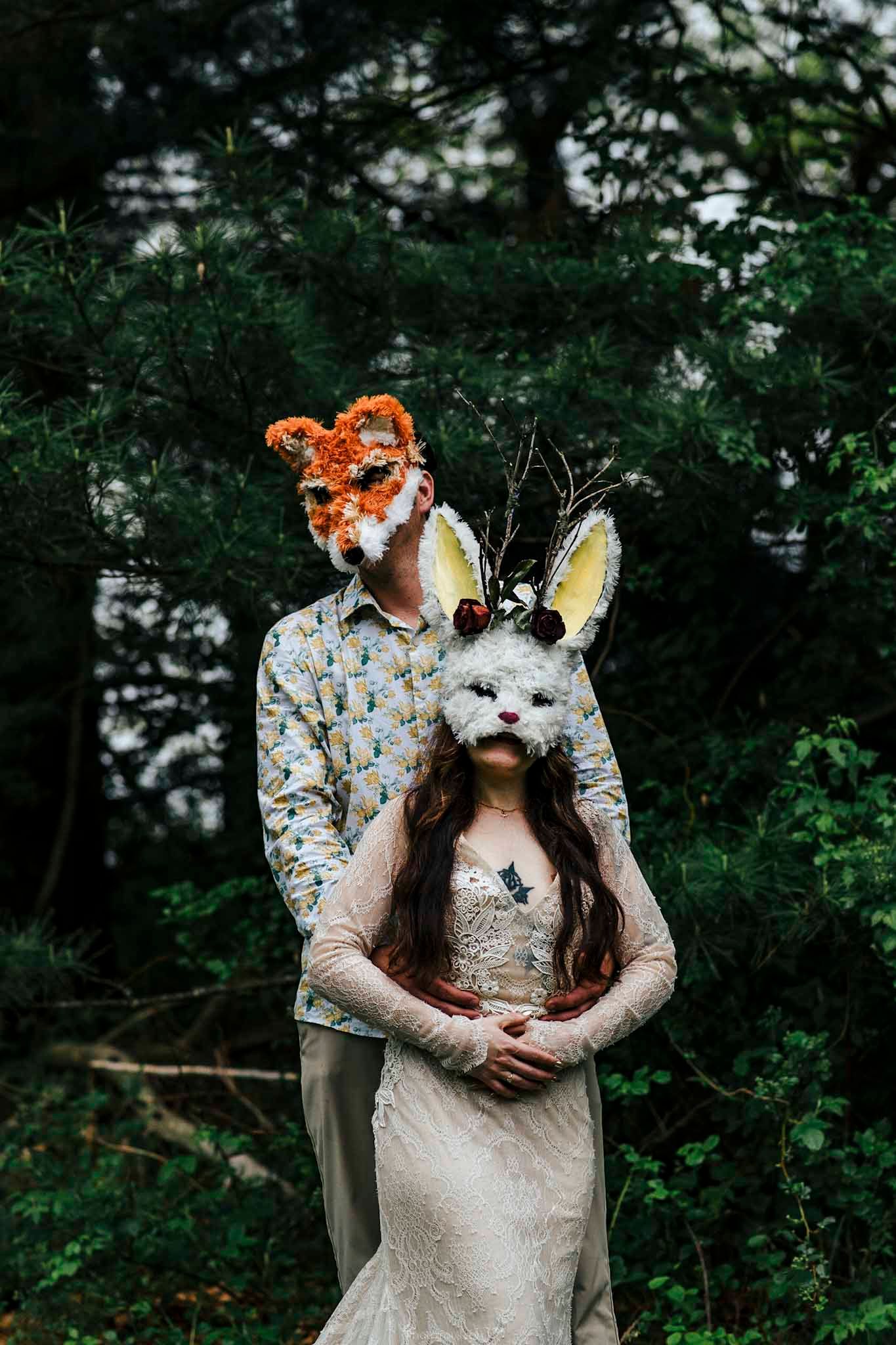 Couple wearing fox and bunny masks posing in a forest, dressed in wedding attire for a whimsical woodland ceremony.