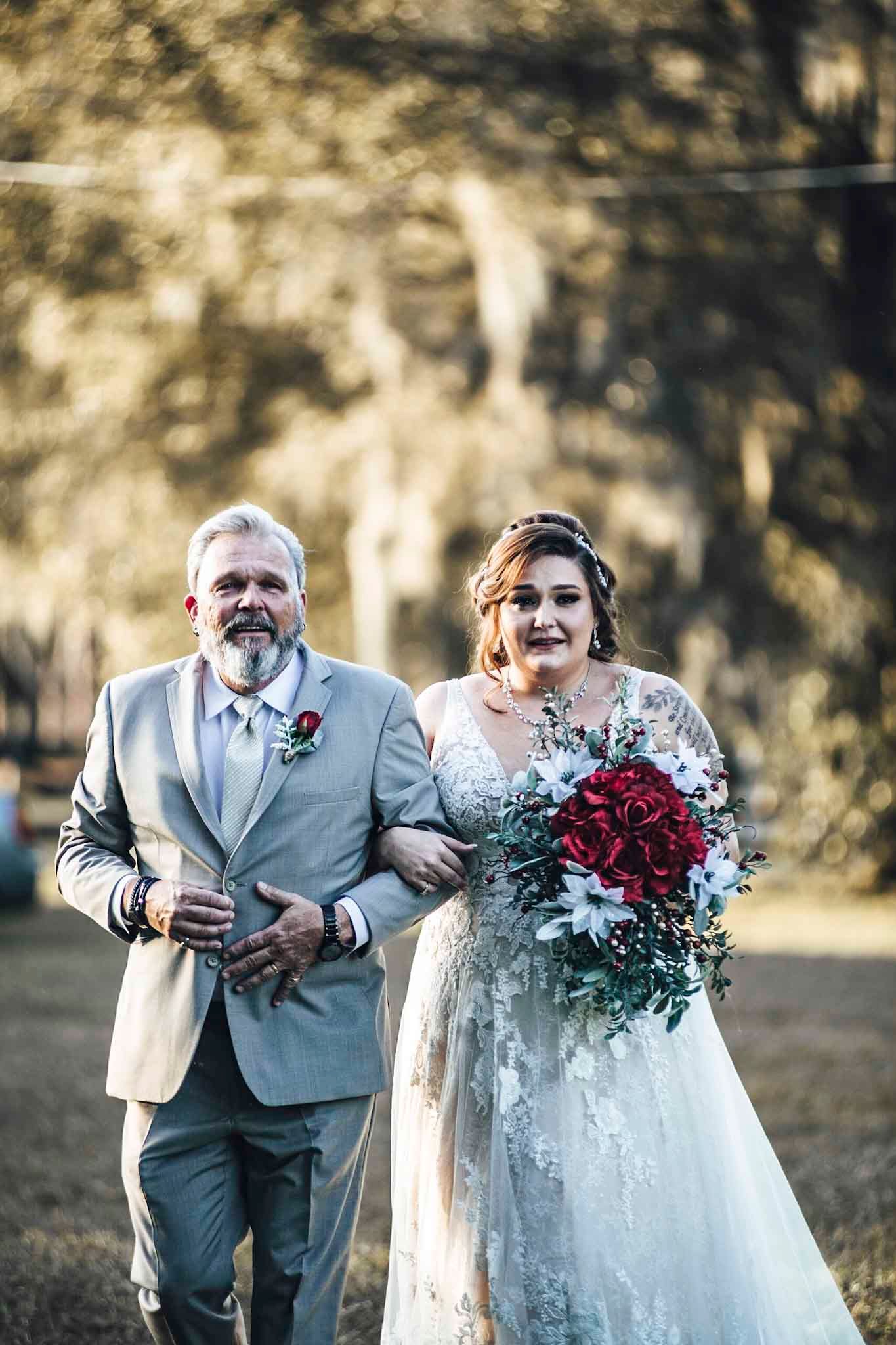 Bride in lace gown holding red rose bouquet walks arm-in-arm with man in gray suit at outdoor wedding ceremony.