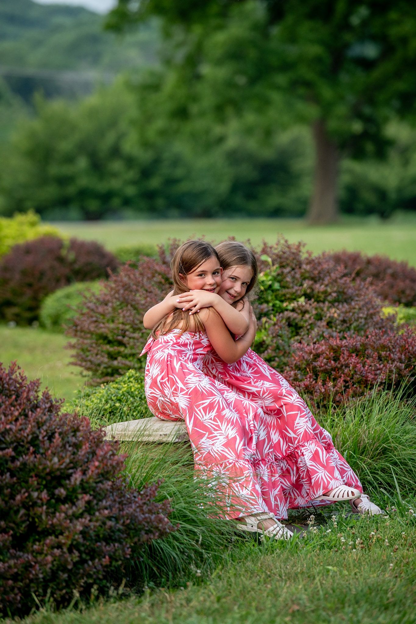 Two young girls in matching pink floral dresses share a warm hug on a garden bench surrounded by lush greenery.