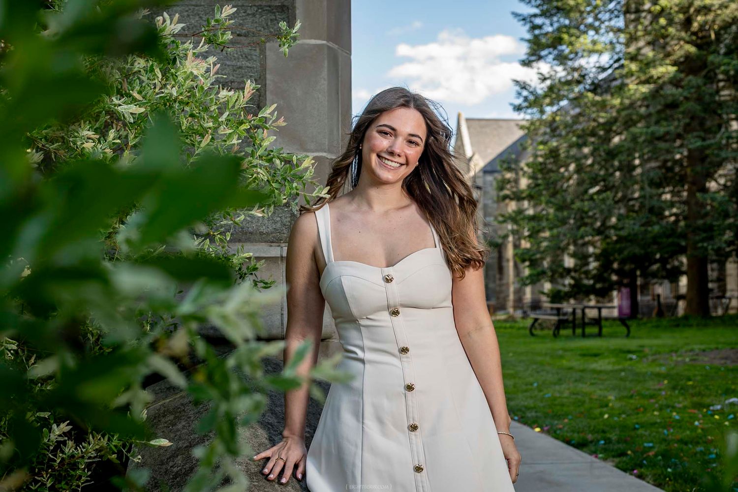 Young woman in white sundress smiling outdoors near stone building and greenery on a sunny day.