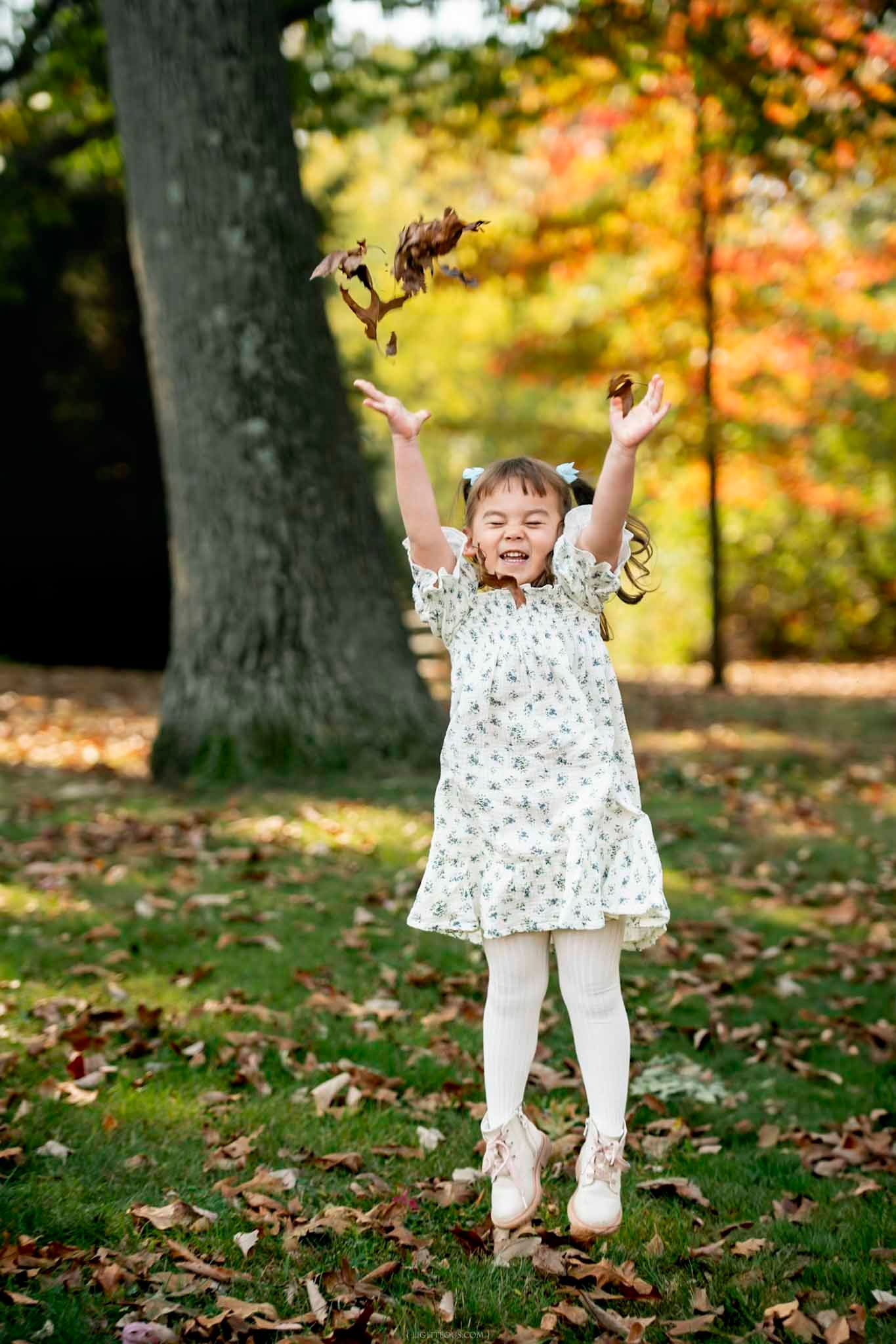 Young girl in floral dress tossing autumn leaves in a park with colorful fall foliage in background.