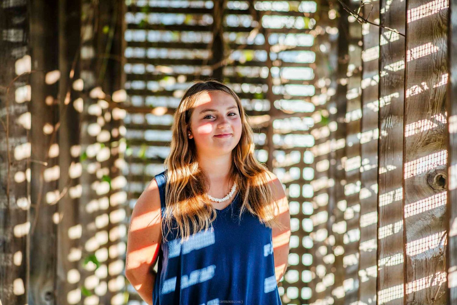 Young woman in a blue dress standing in dappled sunlight between wooden slat walls, looking upward with a gentle smile.