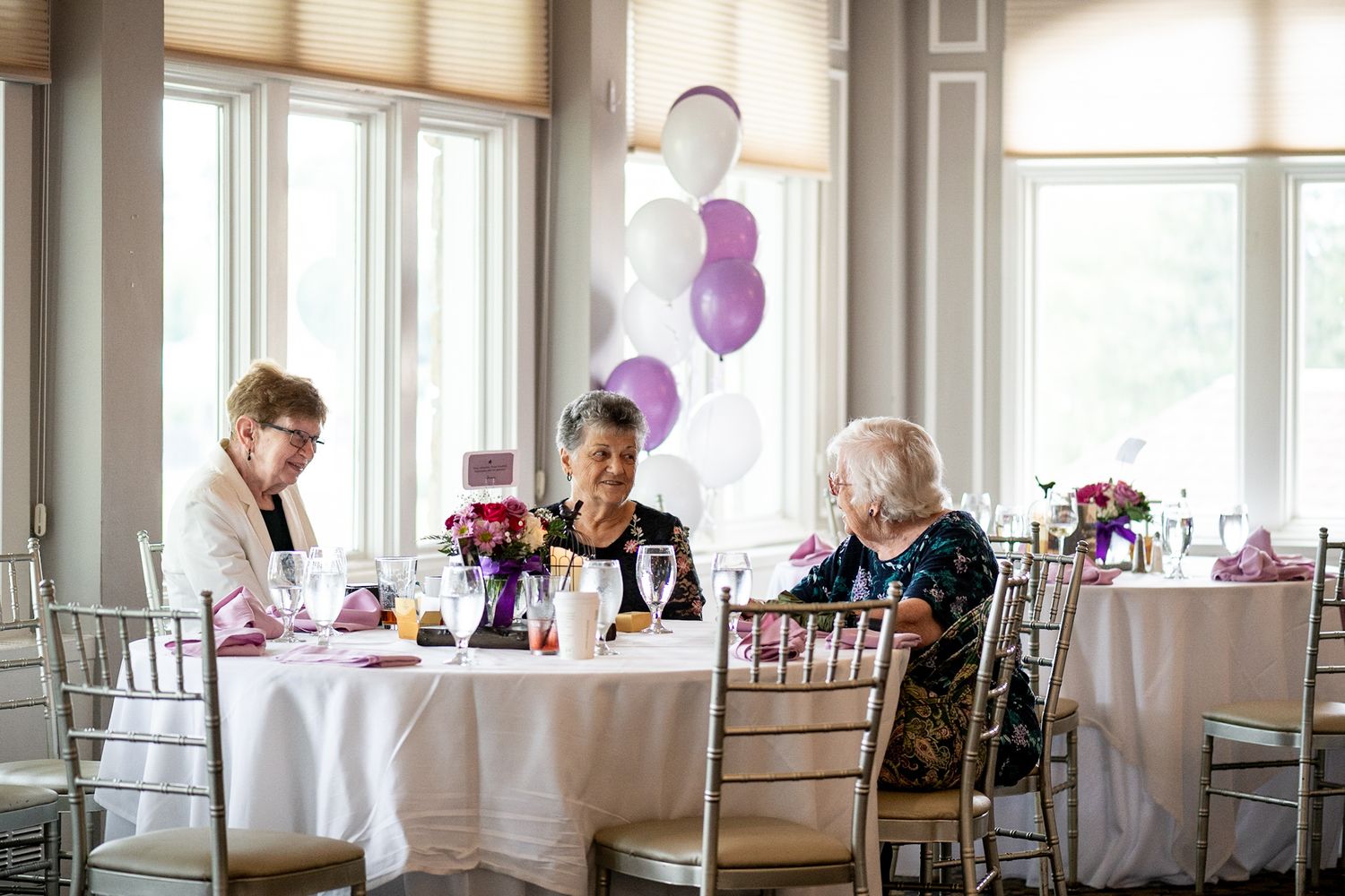 Three elderly women seated at elegantly set tables with purple and white balloon decorations nearby.