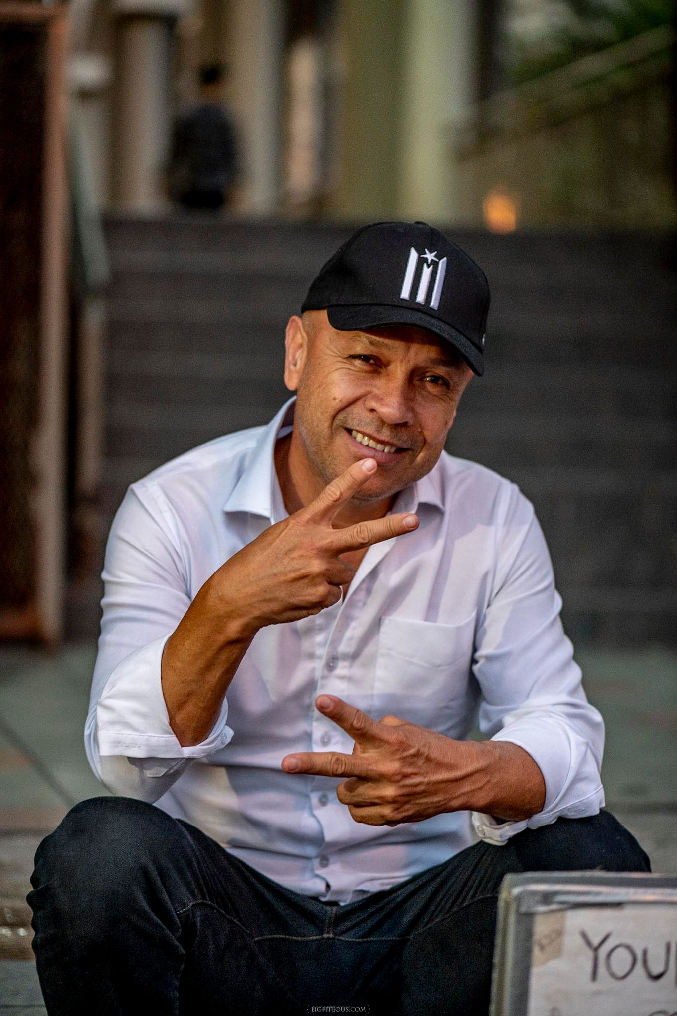 Smiling man in white shirt and black cap sitting outdoors, making hand gestures in a candid street portrait.