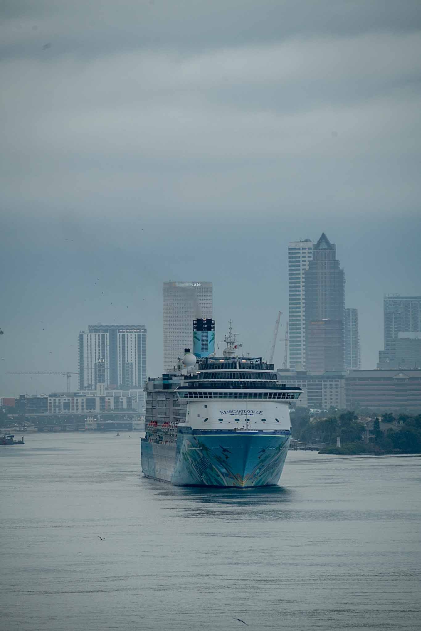 Three large cruise ships sailing in front of a misty city skyline on a cloudy day.