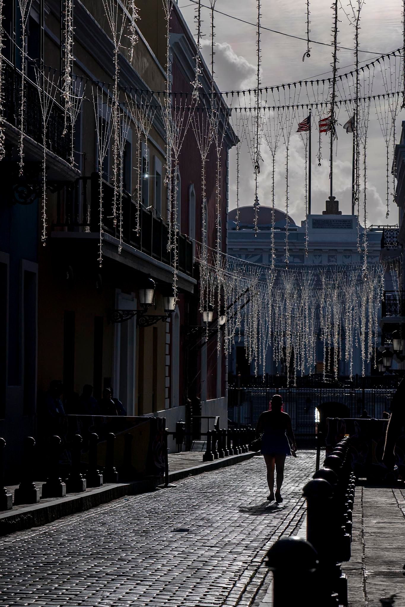 A lone pedestrian walks along a cobblestone street adorned with hanging lights and flags in a historic district.