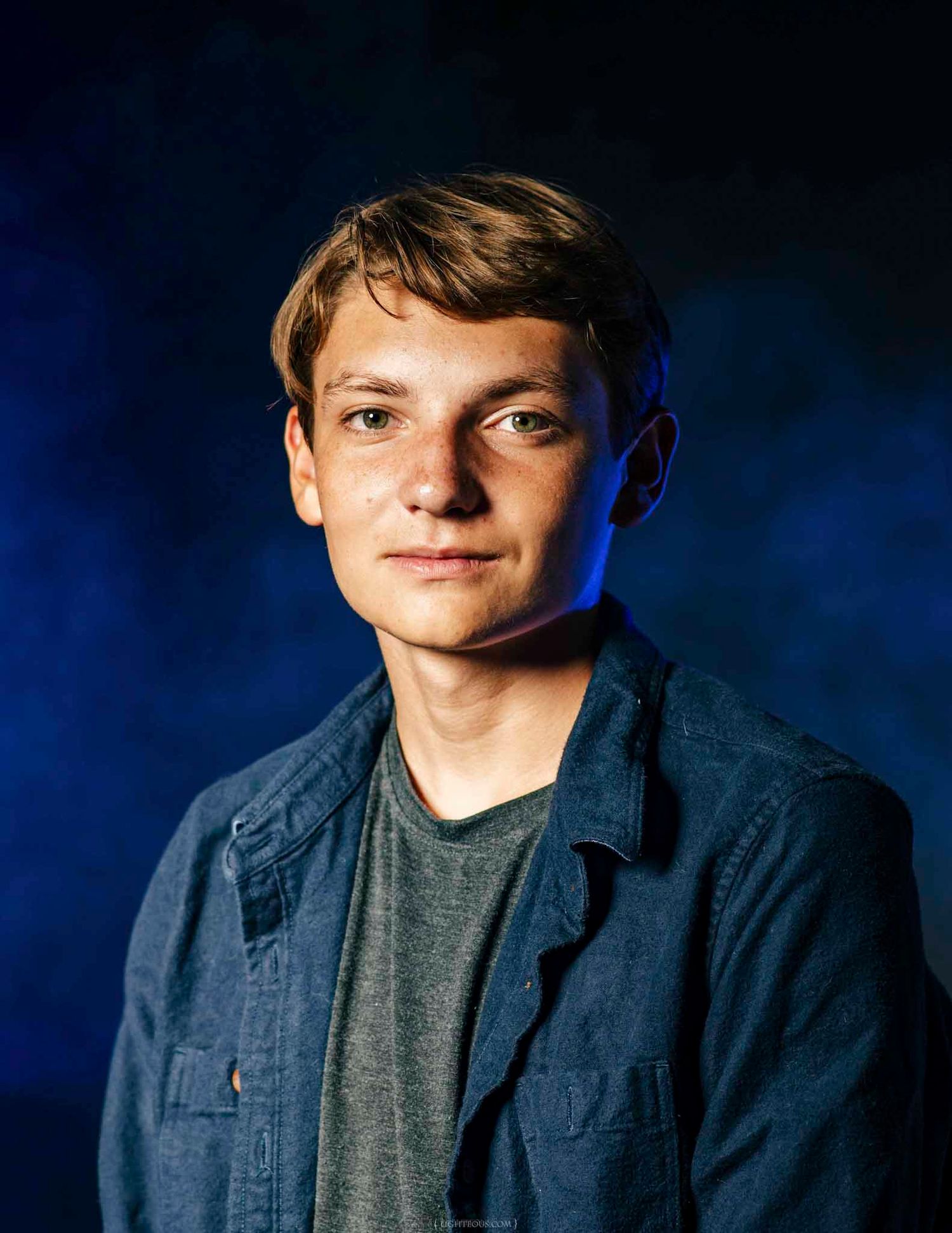 Young man with short brown hair wearing a blue denim jacket, posed against a dark blue studio background.