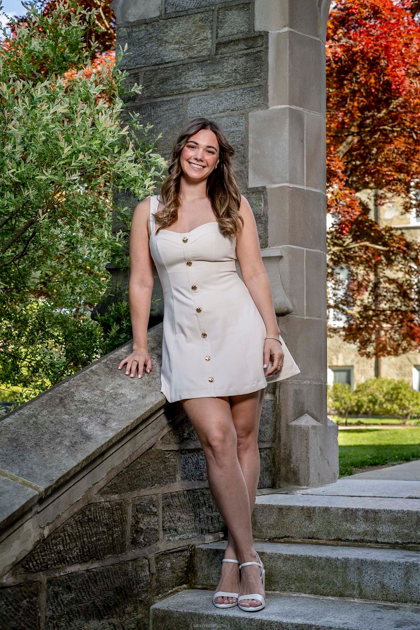 Young woman in beige button-front mini dress poses on stone steps surrounded by autumn foliage.