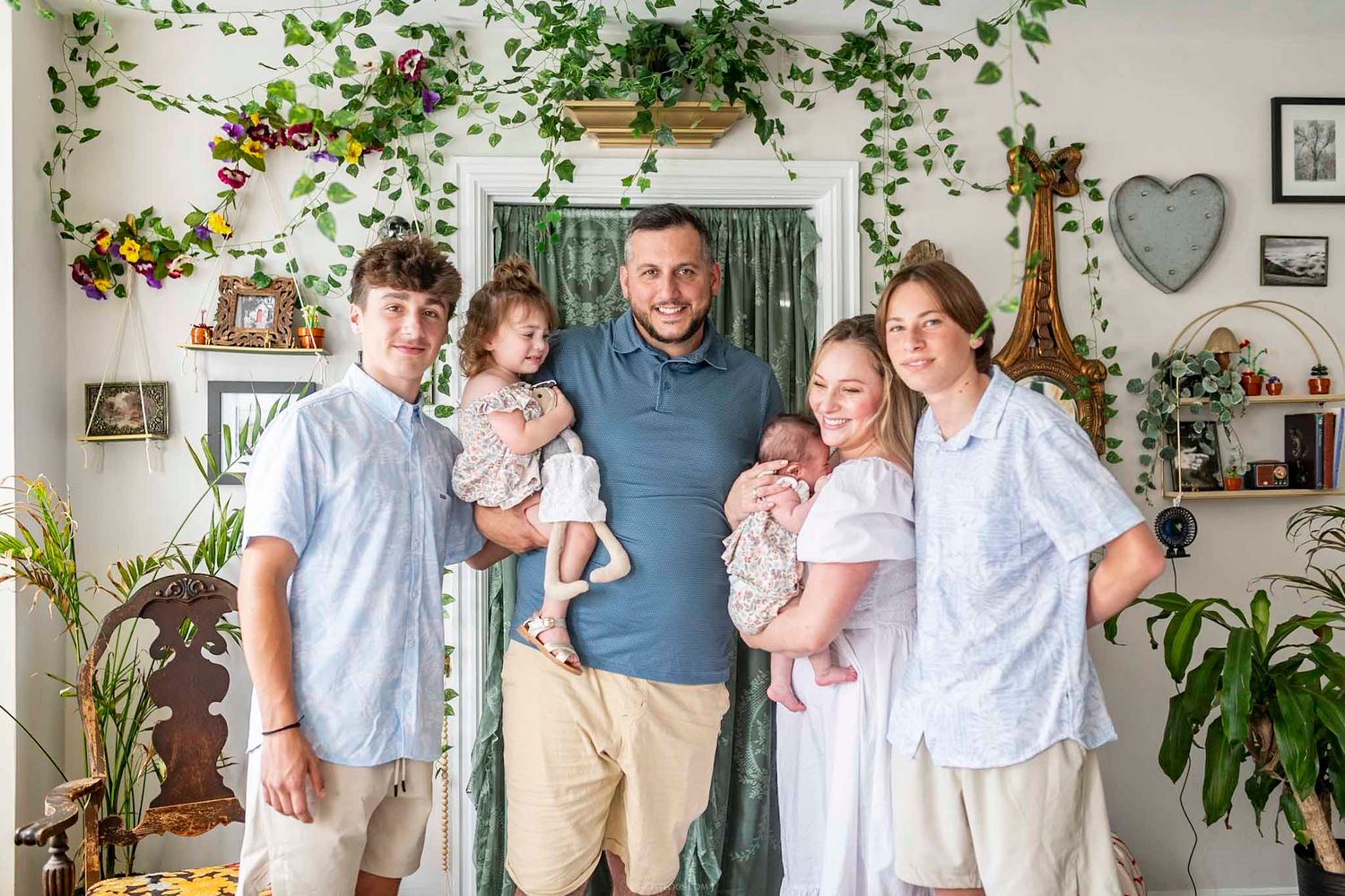 Family of six posing indoors with lush green plants and floral decor, including a newborn baby.