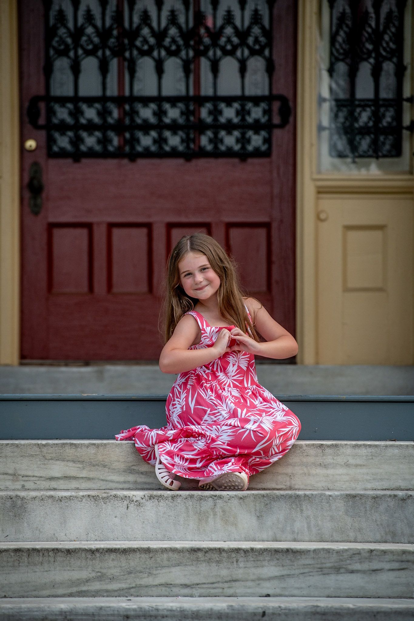 Young girl in pink floral dress sitting on stone steps, smiling and making a heart shape with her hands.