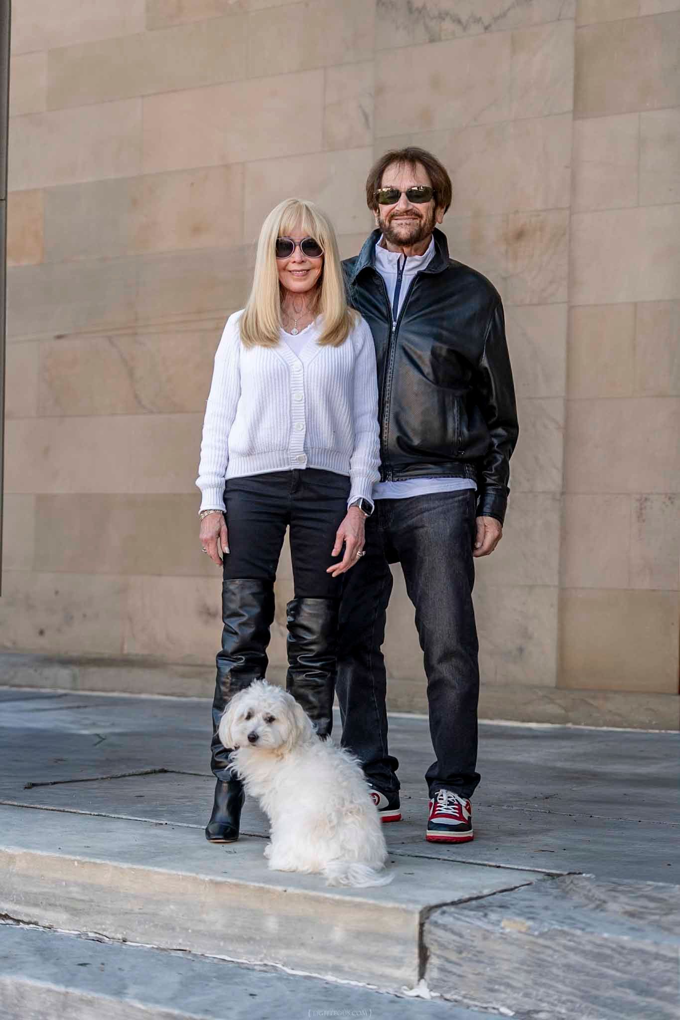 A couple in sunglasses with a white fluffy dog pose outdoors near a stone wall.