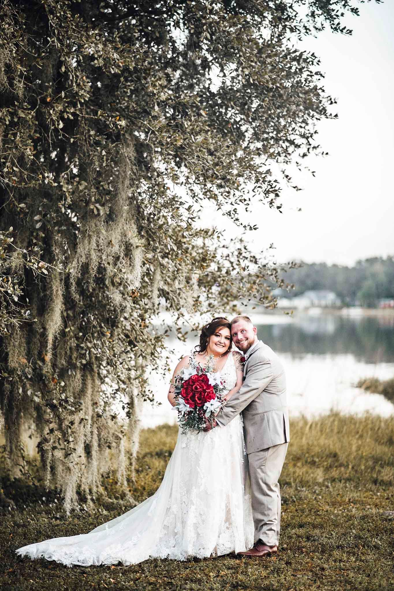 Bride in lace gown holding red bouquet embraces groom in gray suit under moss-draped tree by lakeside.