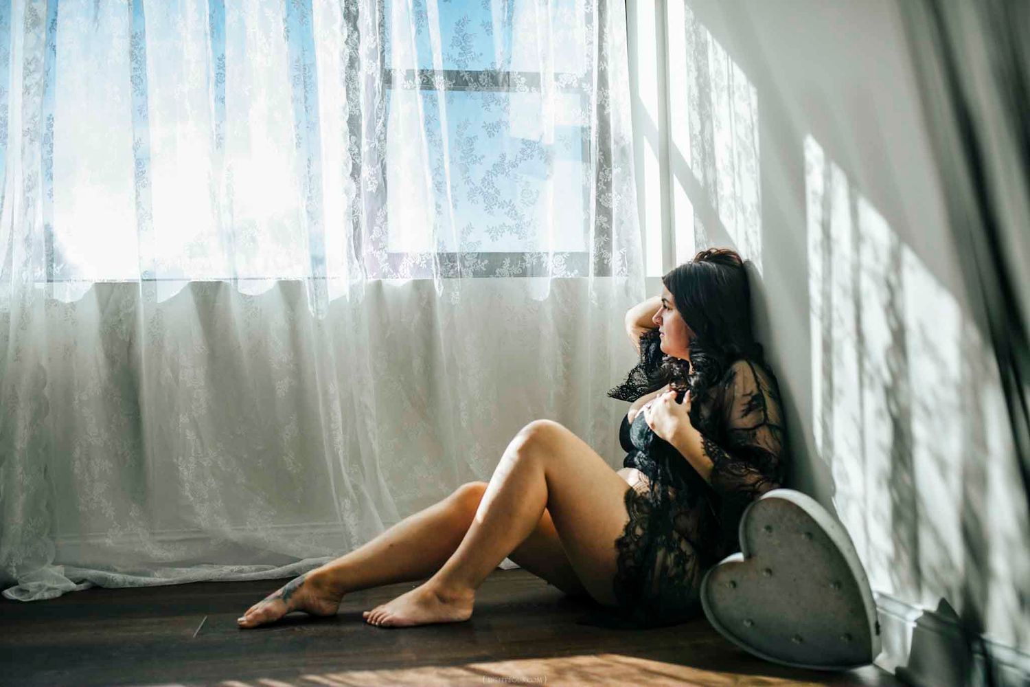 A young woman in a black lace outfit sits on a wooden floor by sheer curtains, gazing thoughtfully into the light.