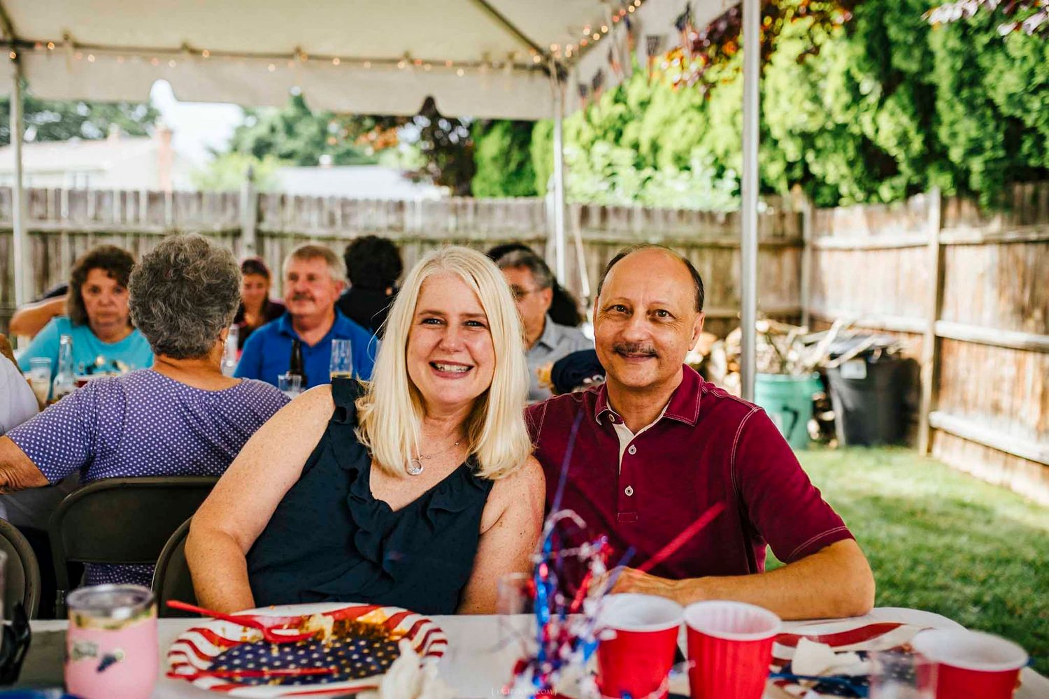 Couple smiling at outdoor Fourth of July backyard party with red cups and patriotic decorations on table.