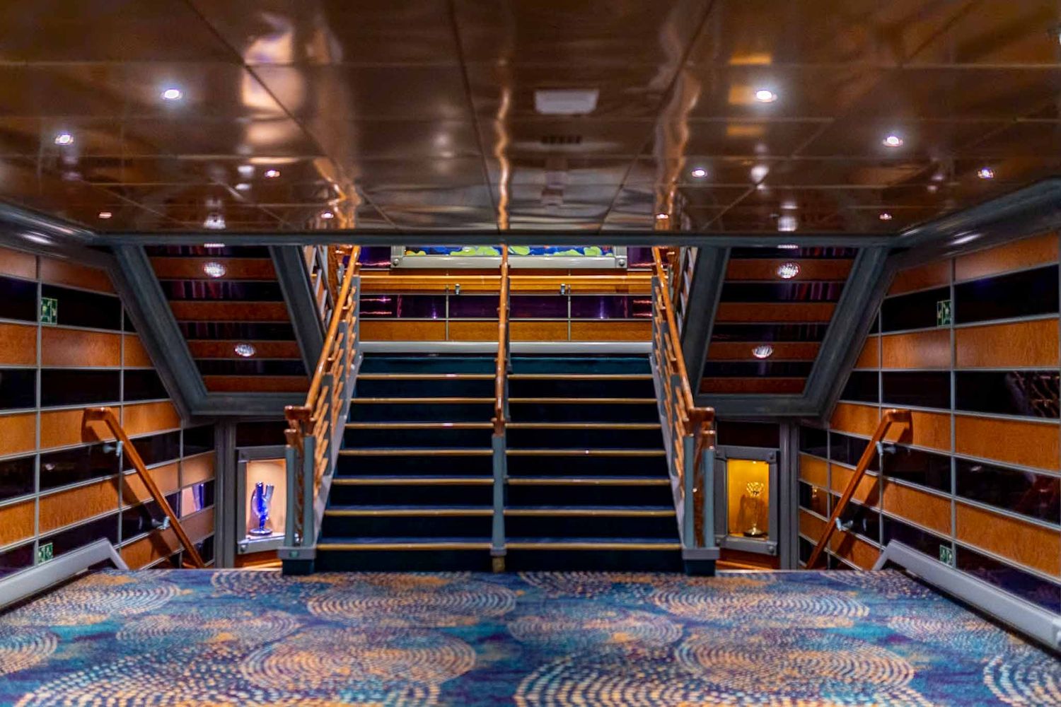 Panoramic view of an empty cruise ship theater with wooden stadium seating and blue carpeted aisles.