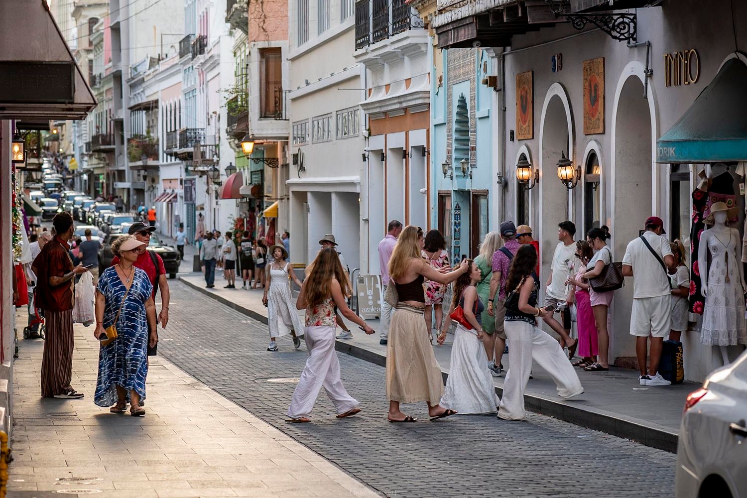 Tourists strolling along a vibrant colonial street lined with white buildings and boutique shops on a sunny day.