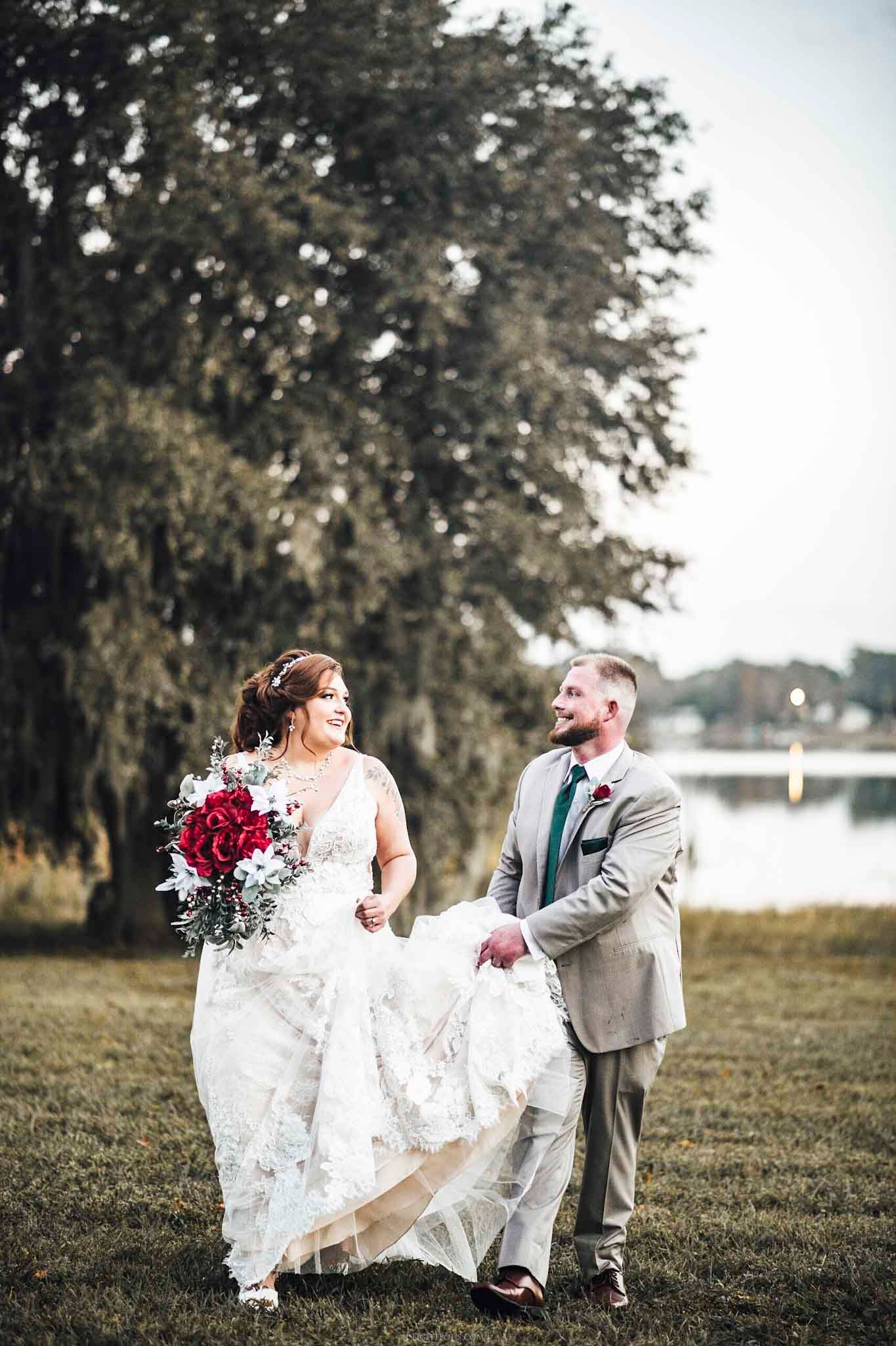 Bride and groom walk hand in hand by a lakeside, she holds red bouquet in white lace gown, he wears a tan suit.