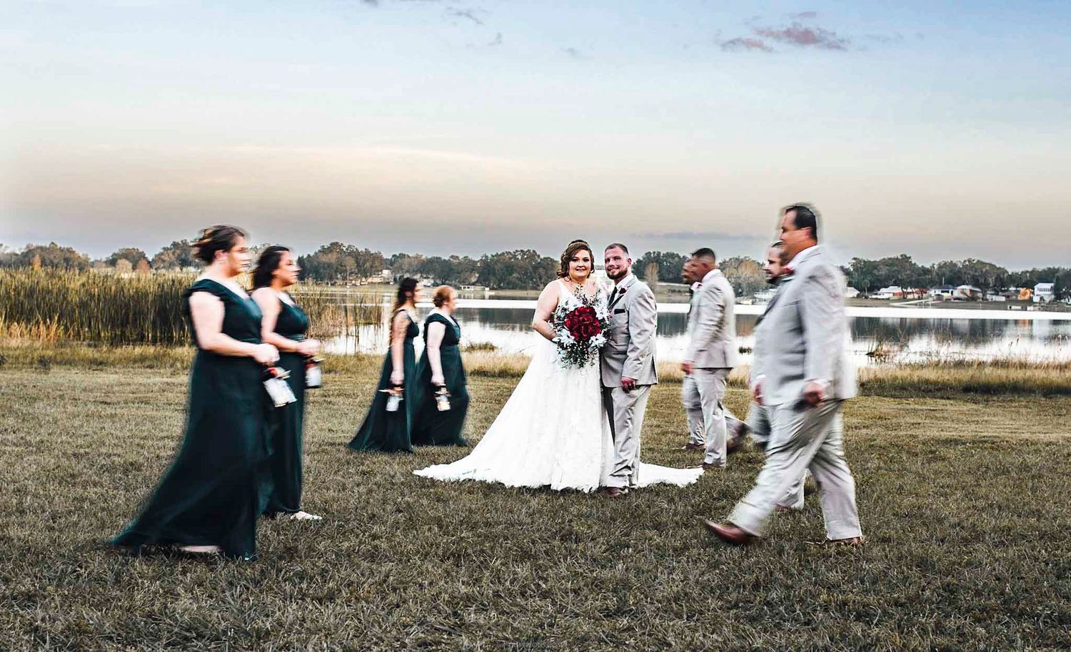 Wedding party walks on grassy waterfront at sunset, bride in white gown with bridesmaids in navy dresses.