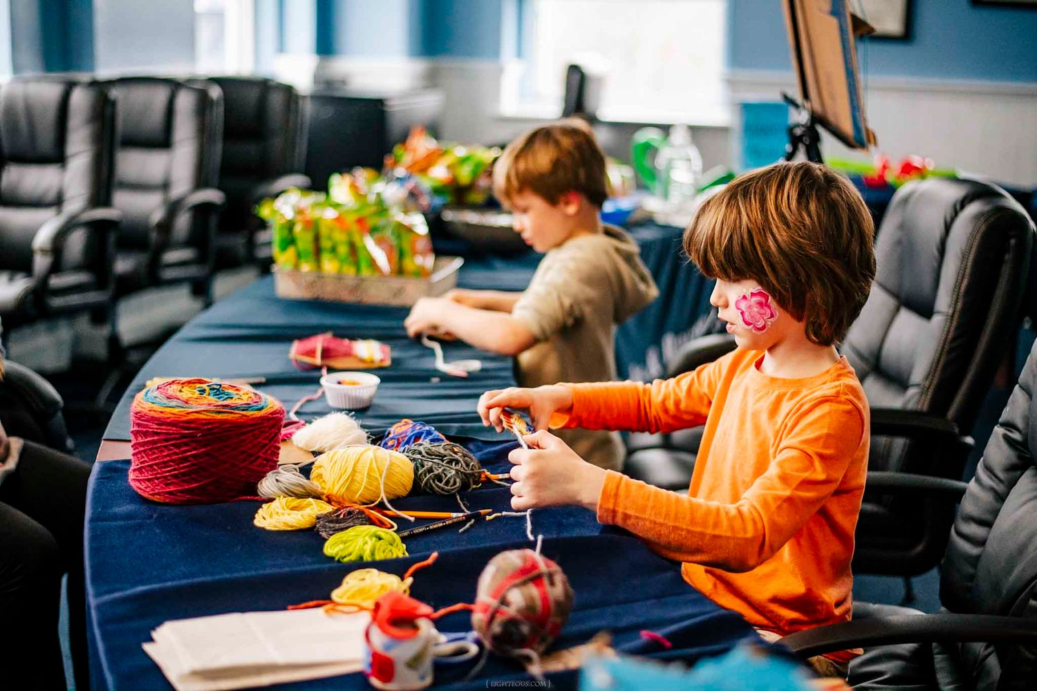 Two children crafting with colorful yarn at a blue table in a classroom setting.