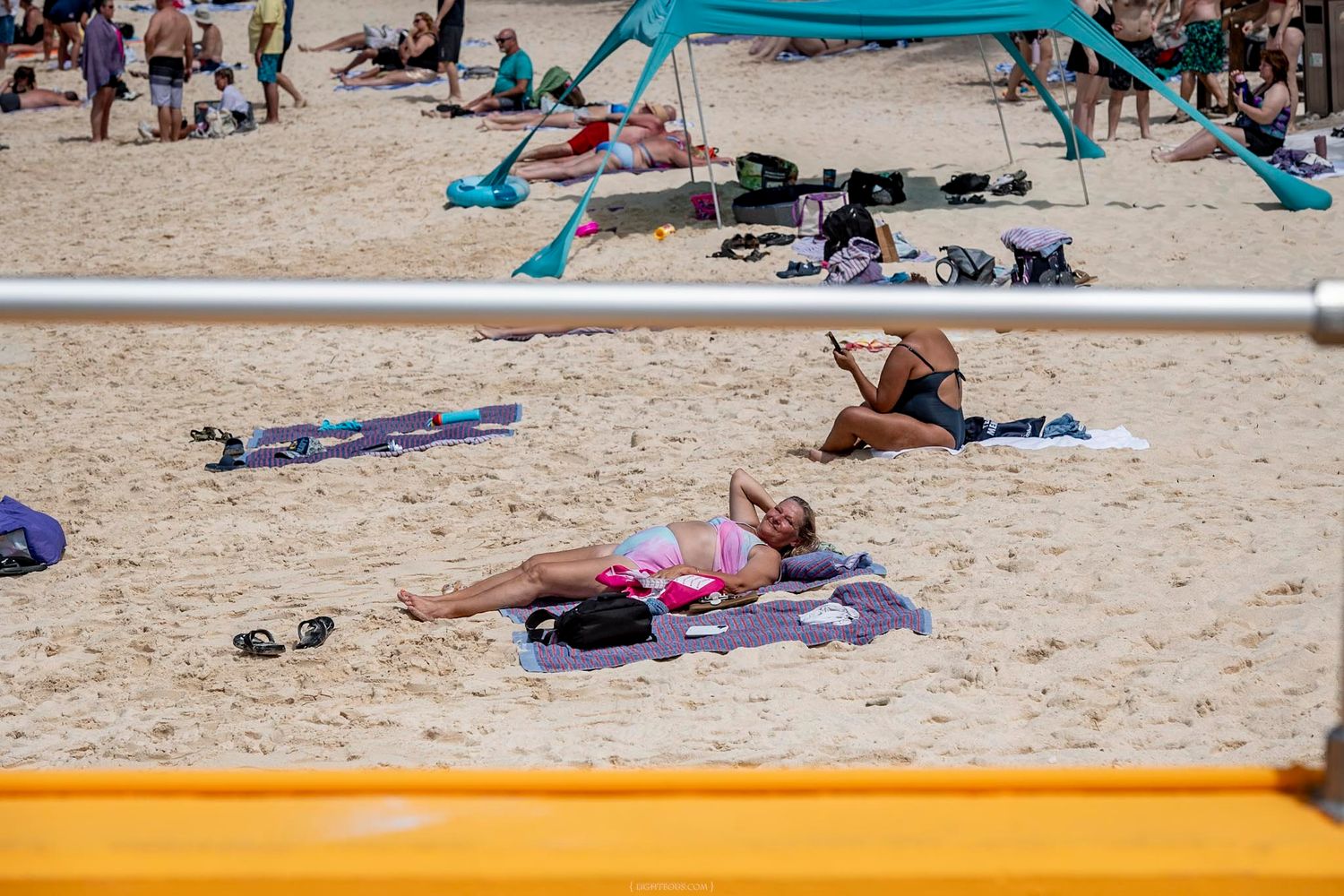 Woman in pink bikini sunbathing on crowded sandy beach near a yellow volleyball net on a sunny day.