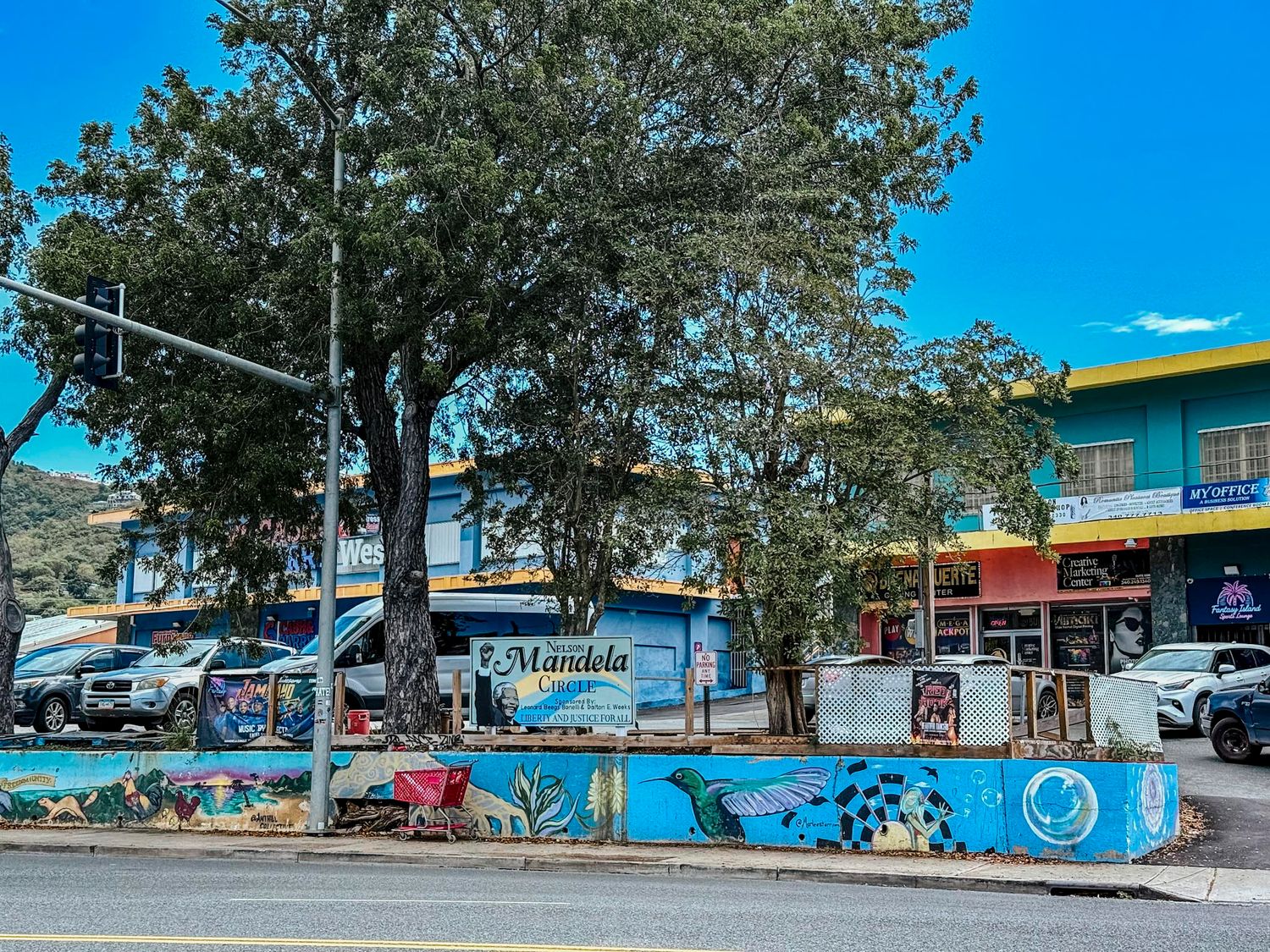 Colorful street scene with a mural-decorated wall, large tree, and shops under a bright blue sky.