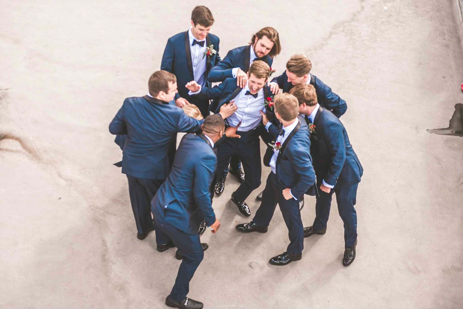 Groomsmen in navy suits playfully surround the groom in a fun wedding party moment captured from above.