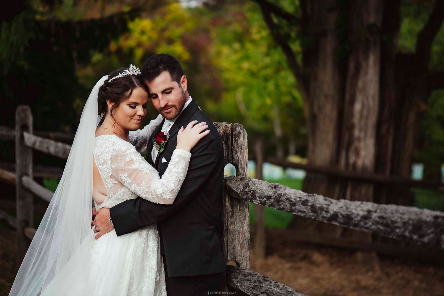Bride and groom embracing near a rustic wooden fence surrounded by lush green trees on their wedding day.