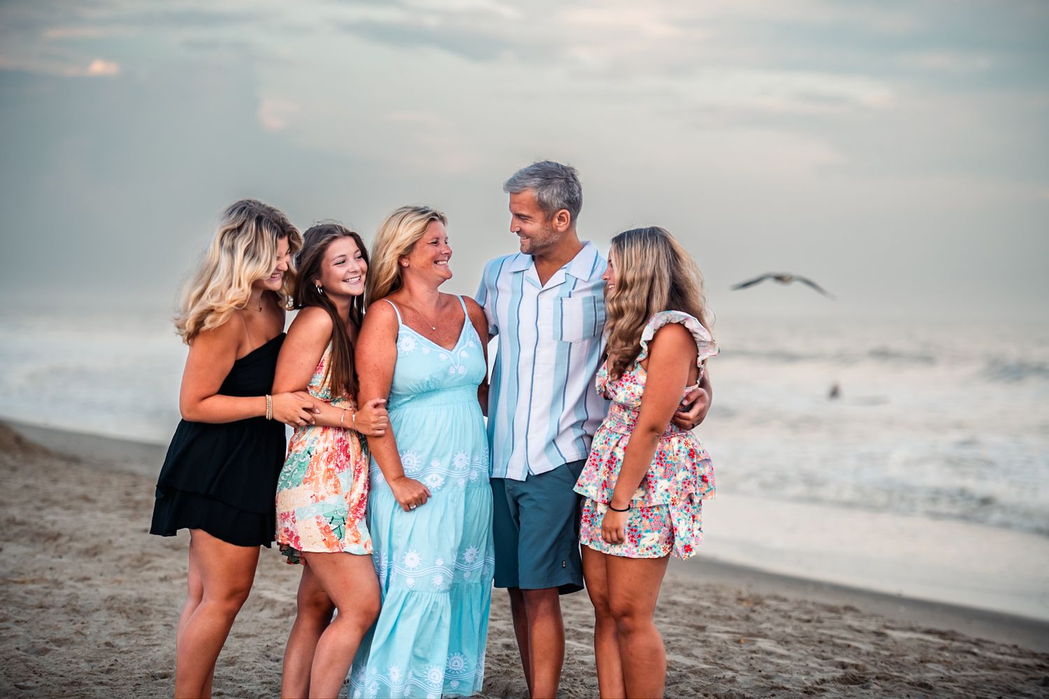 Family of five smiling and embracing on a sandy beach at sunset near ocean waves.