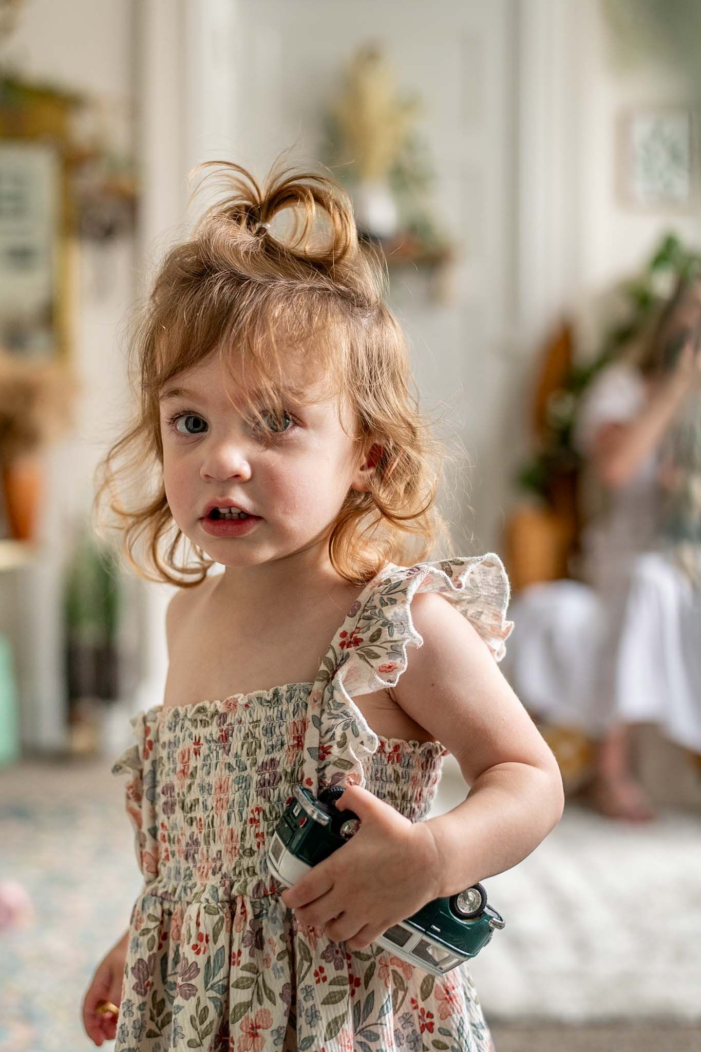 A small child wearing a floral smocked dress holds something while standing in a bright indoor setting.