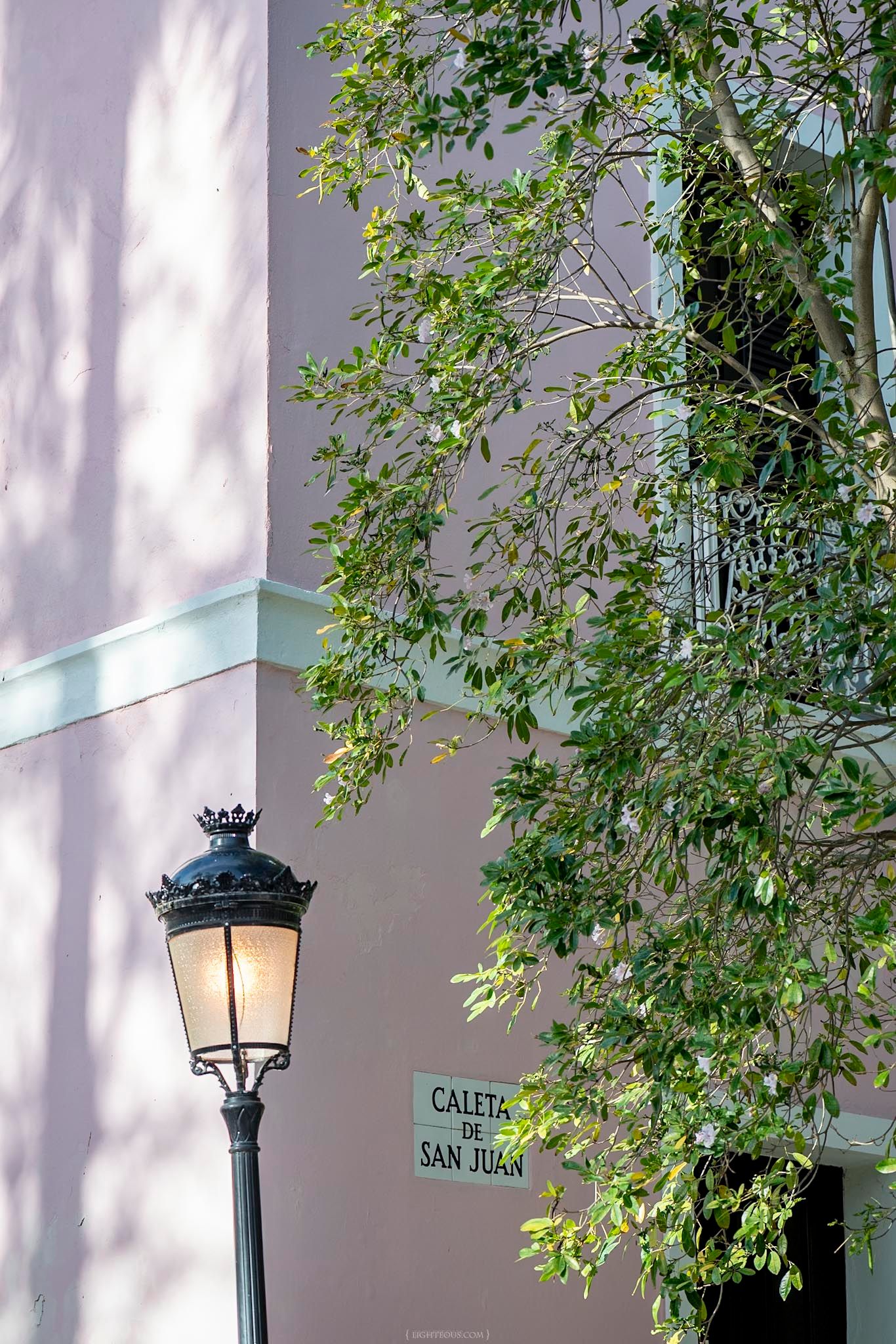 Ornate black street lamp lit up beside a Caleta de San Juan street sign with green trees and pink building.
