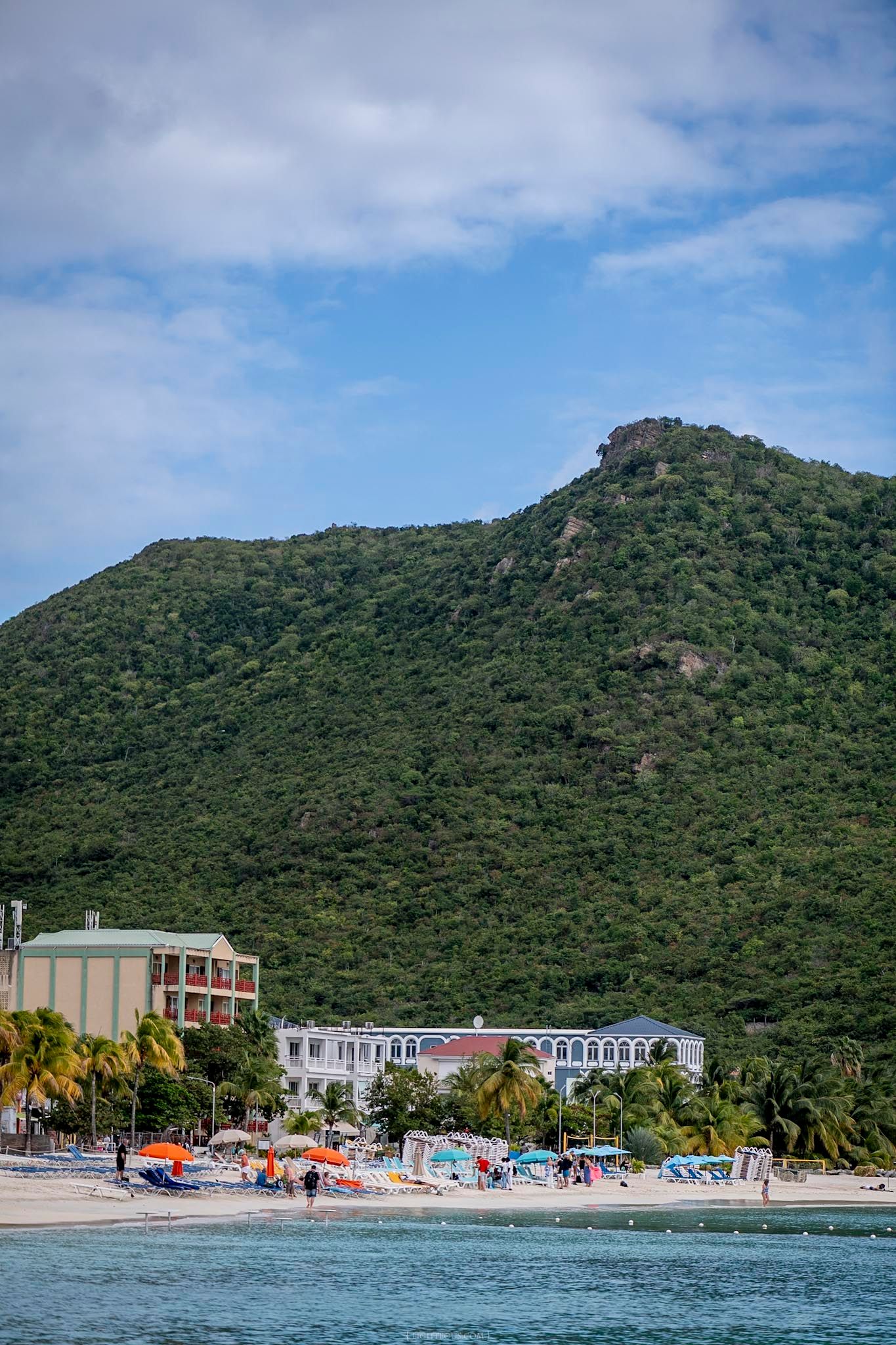 A tropical beach resort with colorful umbrellas, palm trees, and a lush green mountain backdrop under blue skies.