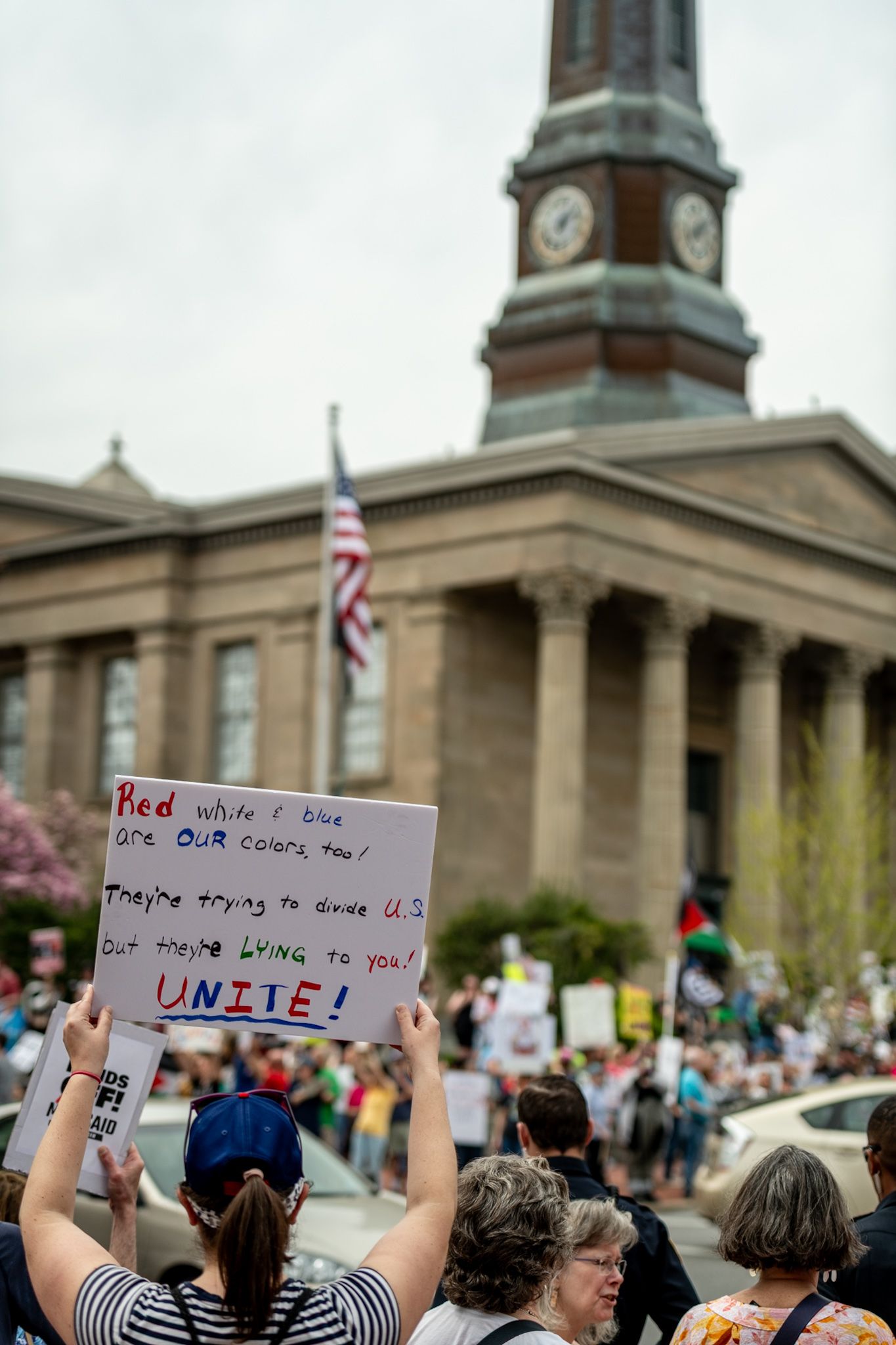 Protesters hold signs in front of a government building with columns and a clock tower during a demonstration.