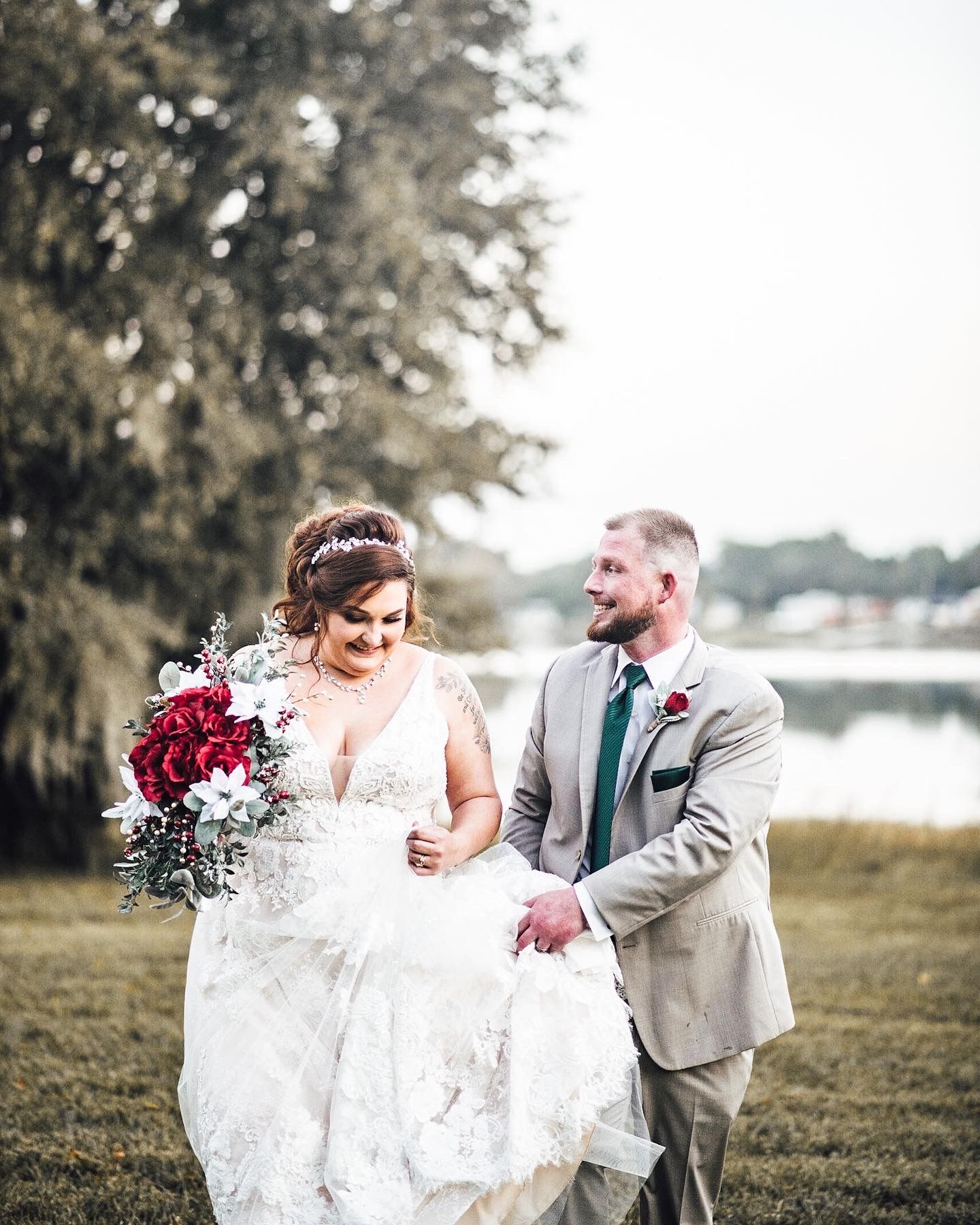 Bride and groom walking outdoors near a lake, bride holding red and white bouquet in lace wedding gown.