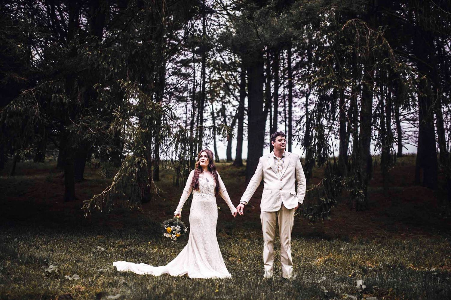 Bride and groom holding hands in a lush forest clearing, bride in white gown, groom in tan suit.