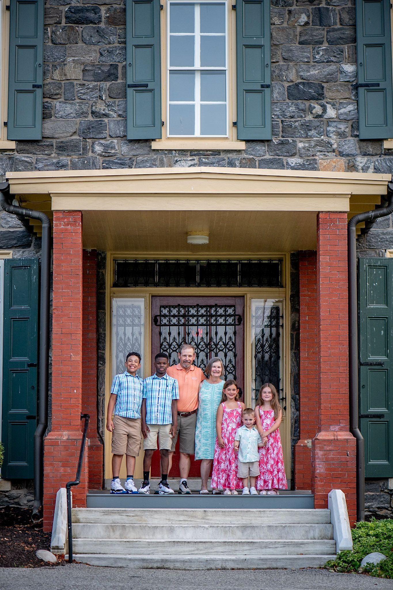 A multigenerational family poses together on the steps of a historic stone building with green shutters.