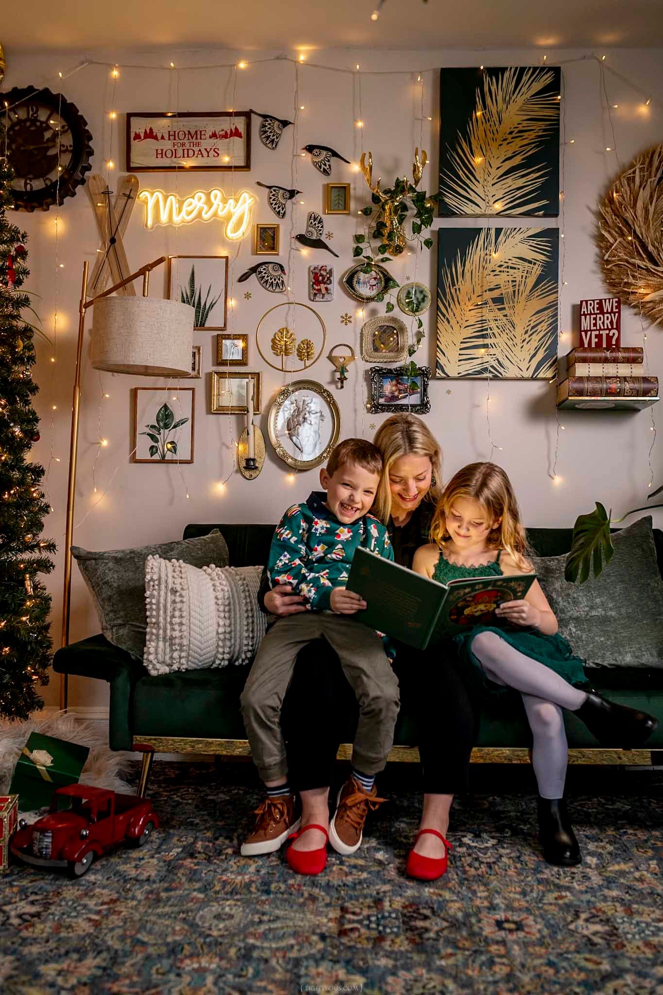 A mother reads a Christmas book with her two children on a green velvet sofa decorated with holiday lights and festive wall decor.