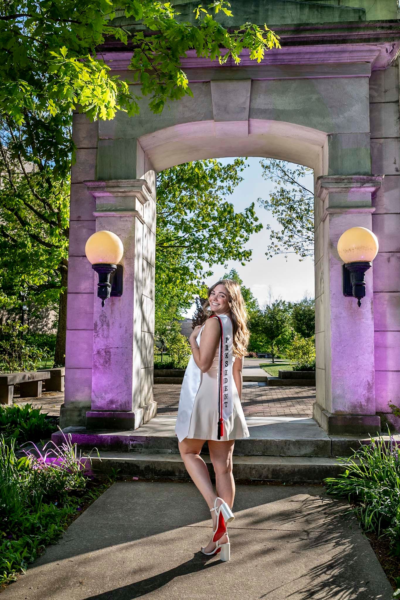 Young woman in white dress poses under illuminated stone arch surrounded by lush green trees in a park.